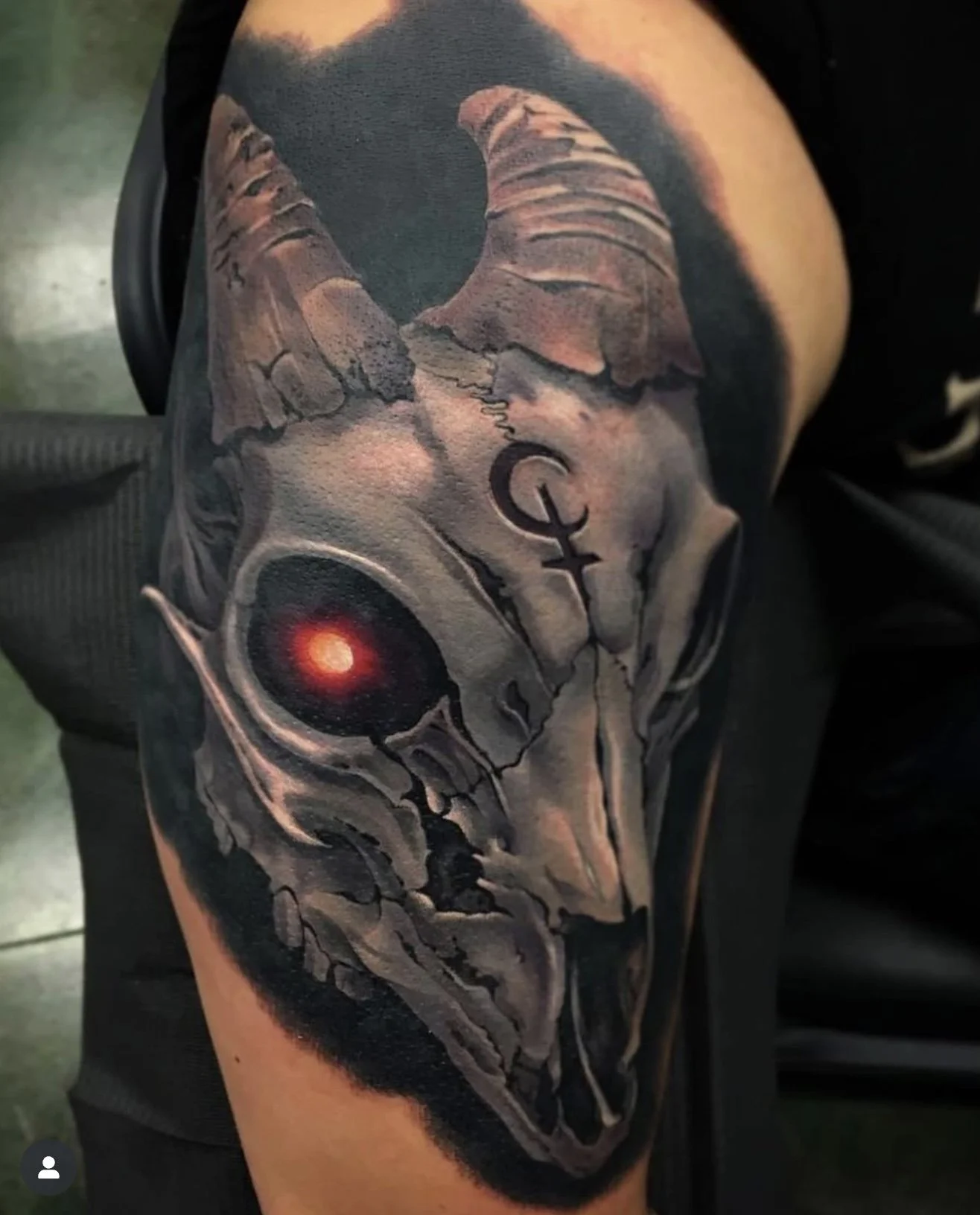 A realistic tattoo of a white skull with a red eye and a horned skull design, covering the person's upper arm.