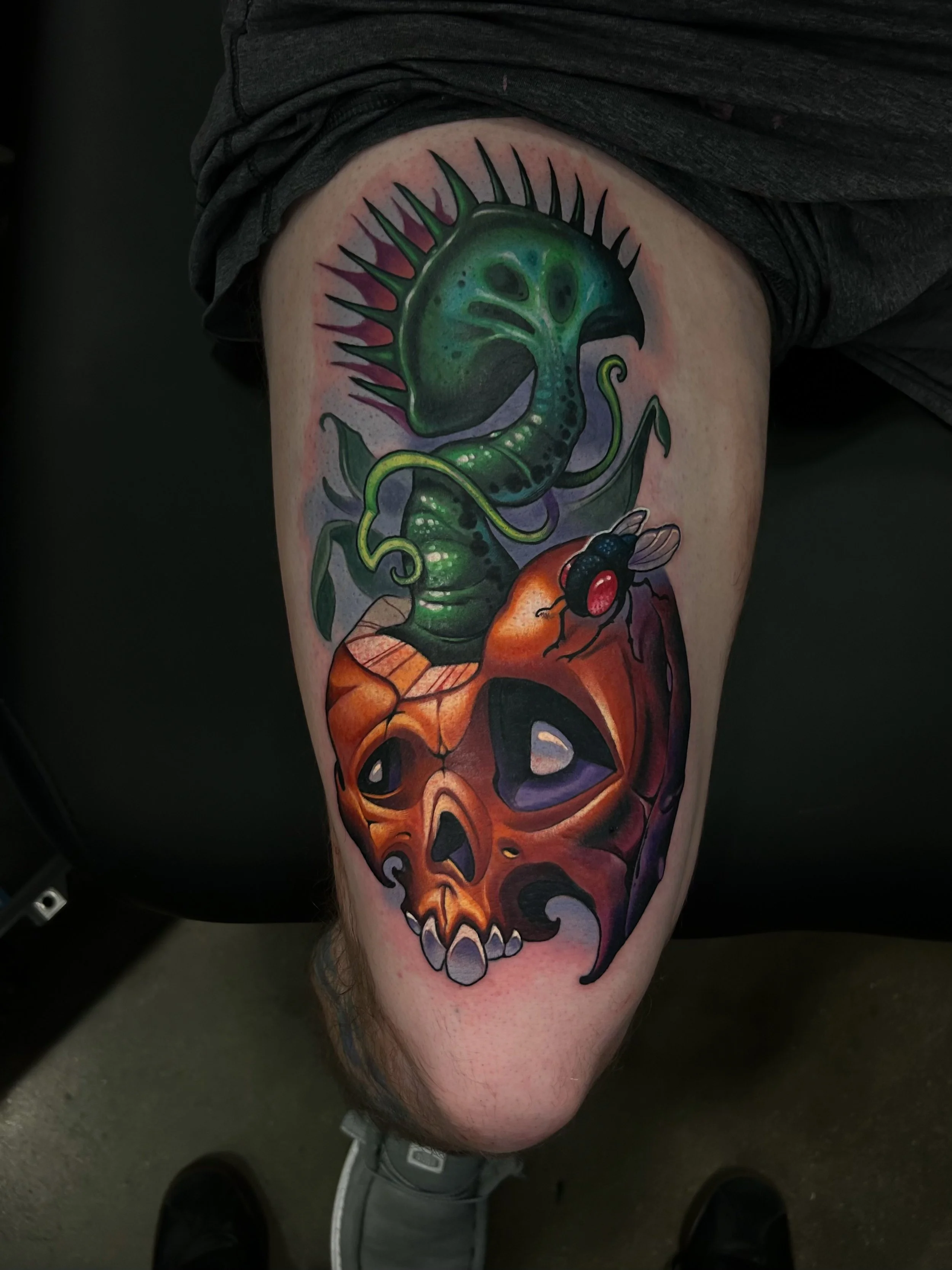 Colorful tattoo of a green dinosaur with spikes on its back, perched on a human skull with a fly on it, on a person's thigh.