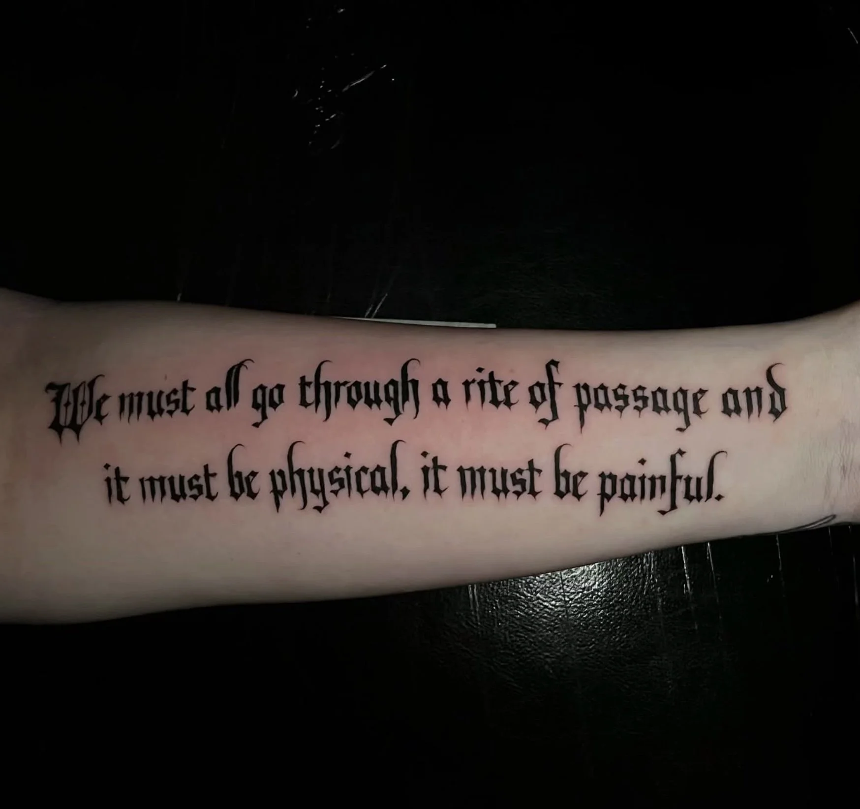A person's arm with a tattoo of a quote written in black gothic-style font.