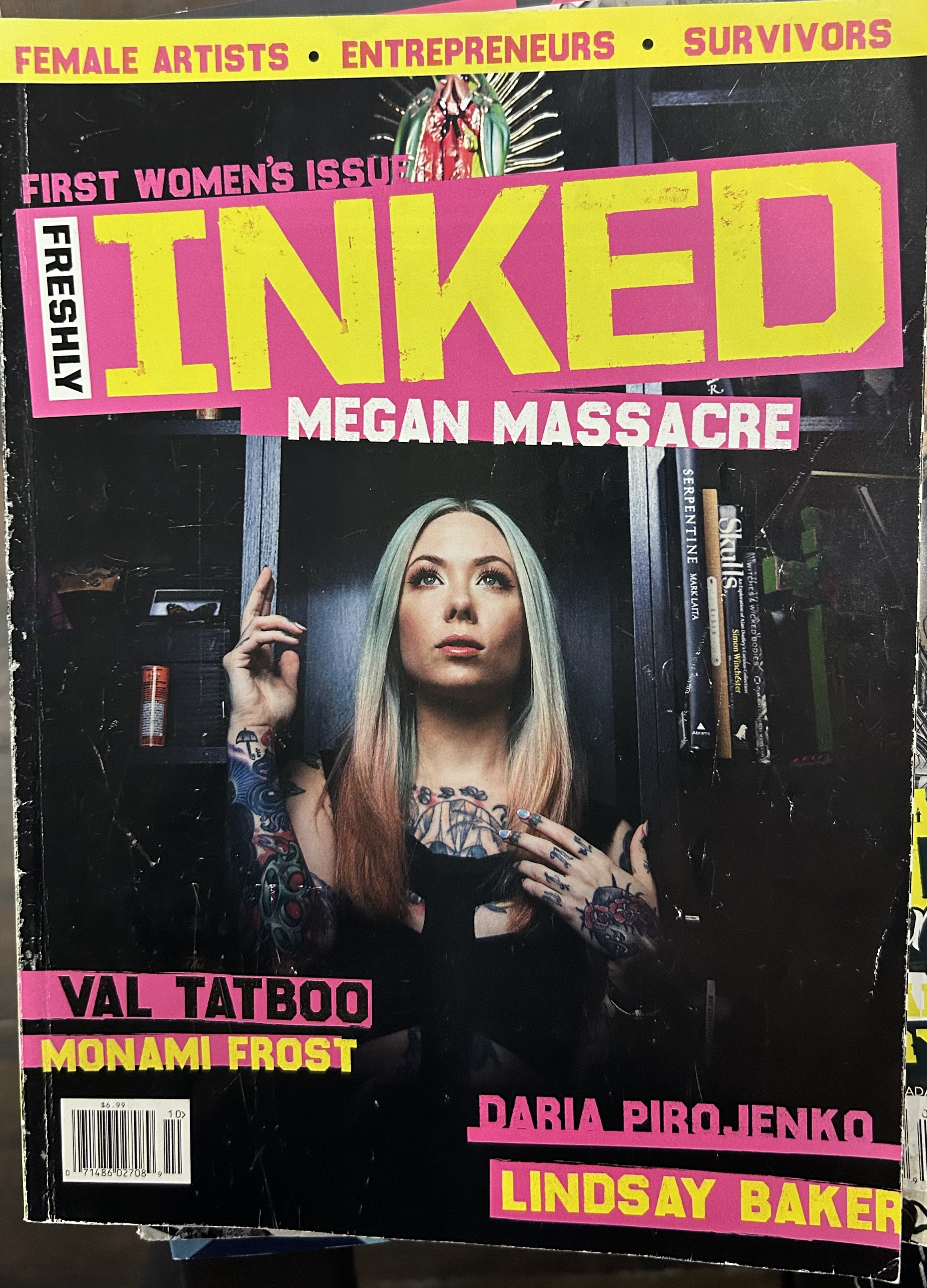 Cover of a magazine titled "INVOKED Megan Massacre" featuring a woman with dyed green and pink hair, tattoos, and makeup, standing in front of bookshelves. The cover highlights female artists, entrepreneurs, and survivors, with other featured names listed such as Val Tattoo, Monami Frost, Daria Pirojenko, and Lindsay Baker.