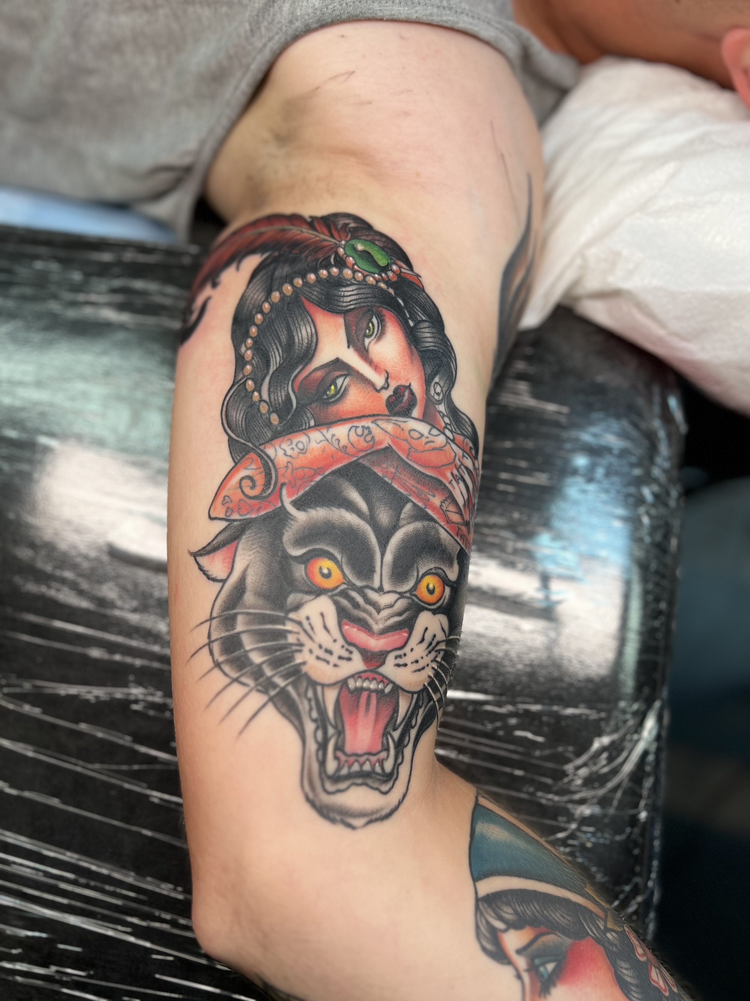 Tattoo of a woman's face with dark hair, green eyes, and red makeup, surrounded by decorative elements on upper arm.