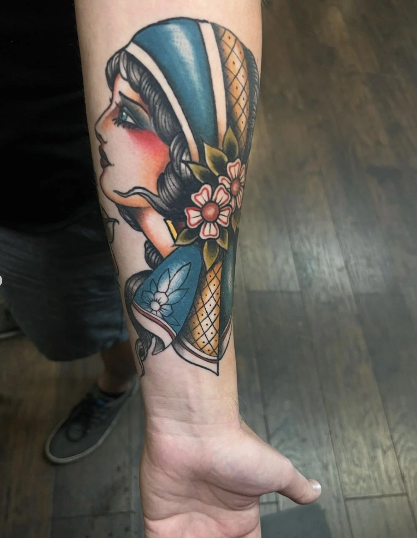 Tattoo of a woman's profile with dark hair, blue headscarf, flowers, and traditional decorative elements on the forearm.