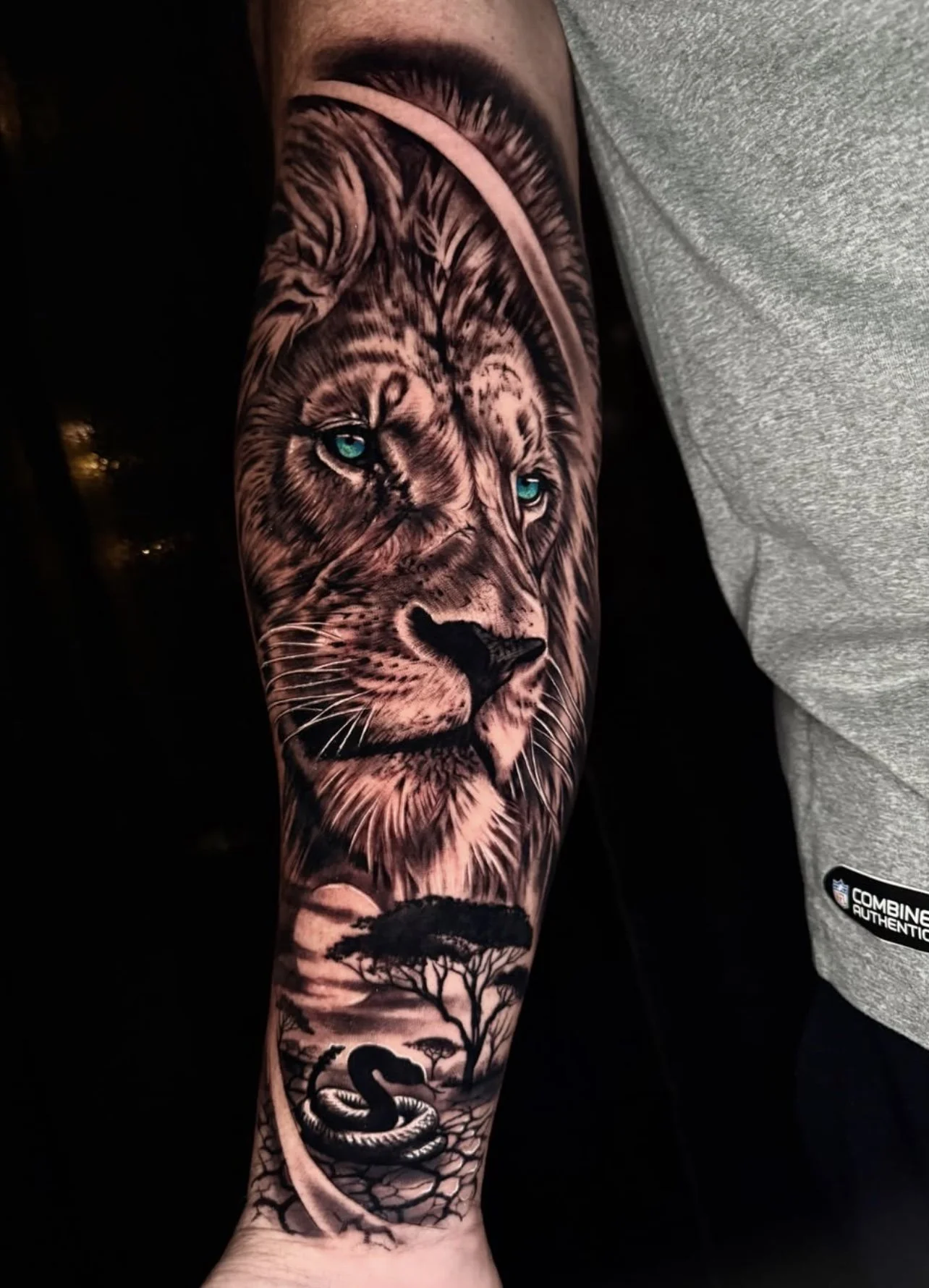 Tattoo of a lion's face with piercing blue eyes, Africa landscape, acacia trees, and a snake on a person's arm.