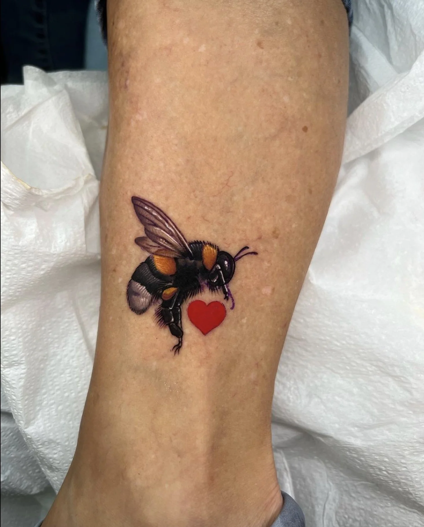 Tattoo of a bee with orange and black colors holding a red heart on a person's arm.