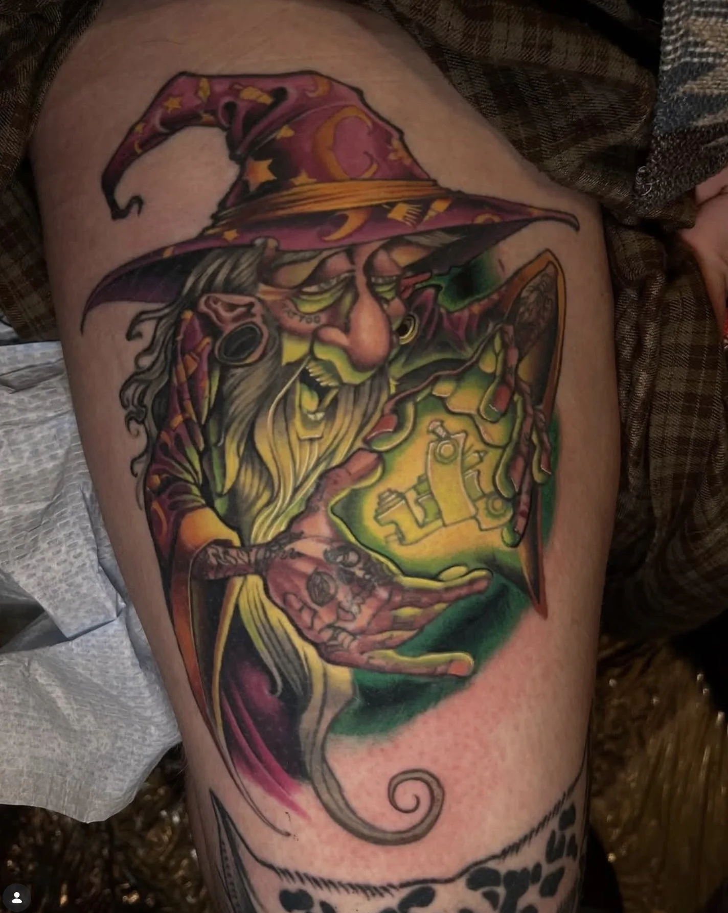 Colorful tattoo of a witch with a pointed hat and long gray hair, holding a broomstick, on a person's upper arm.