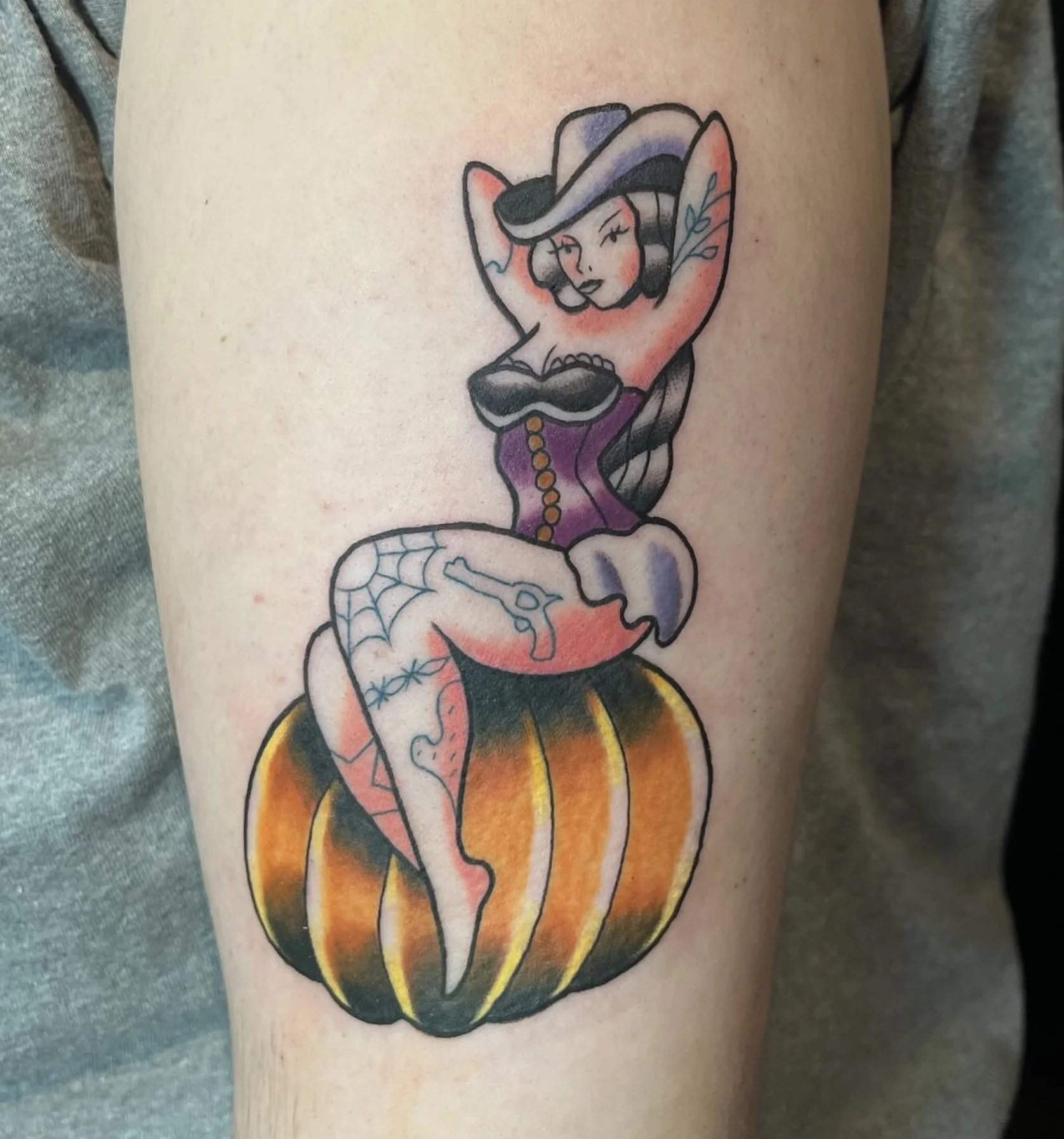 Tattoo of a vintage pin-up girl with a cowboy hat, sitting on a pumpkin, surrounded by a spider web.