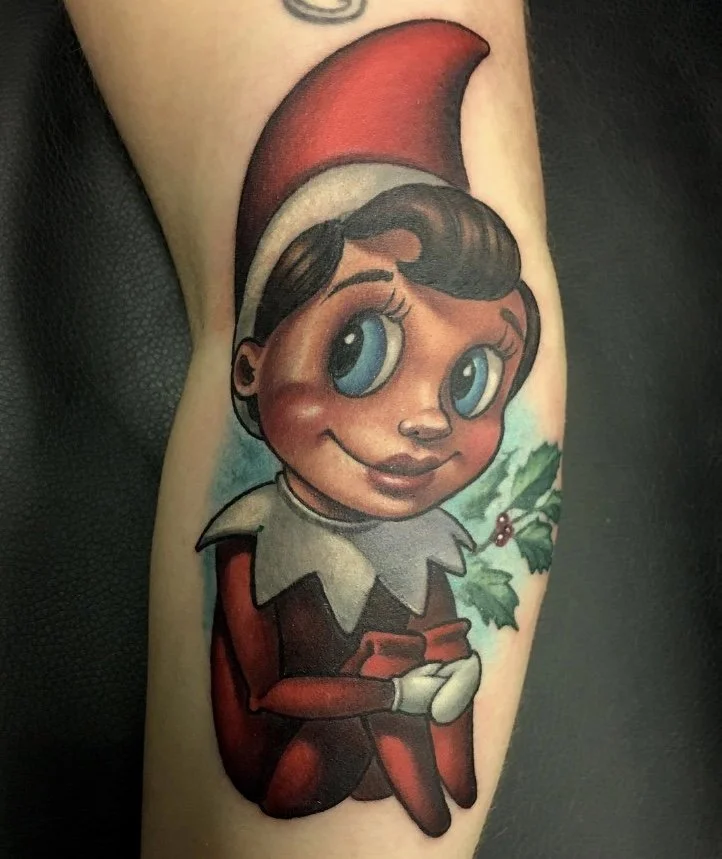 Colorful tattoo of a cartoon elf girl with big blue eyes, wearing a red and white Christmas hat, red dress, white gloves, and sitting next to holly leaves with berries.