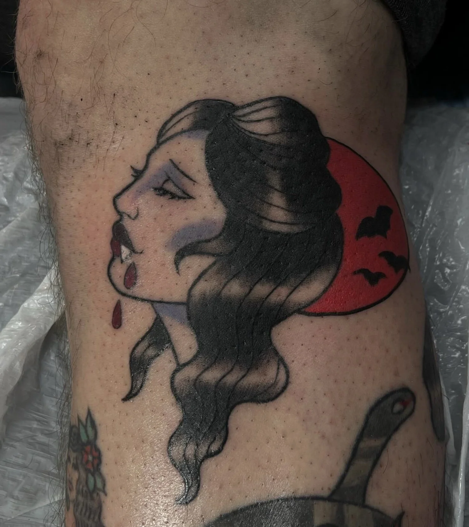 Tattoo of a woman with long flowing hair, closed eyes, and dark lipstick, with a red moon behind her and bats flying in the background.