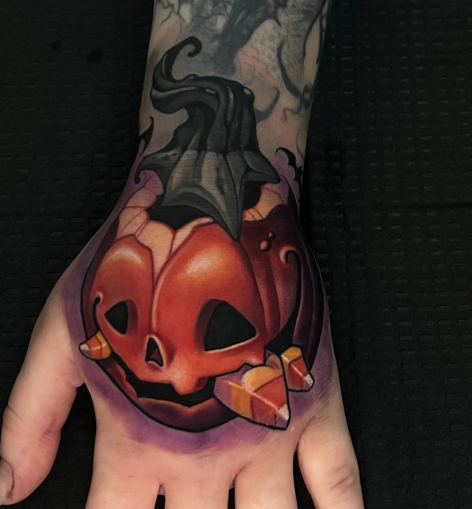 Colored tattoo of a jack-o'-lantern pumpkin with a carved face, accompanied by a bat-shaped leaf, on a person's hand and wrist.