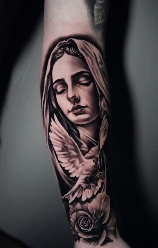 Black and gray tattoo of a woman's face with eyes closed, a dove in front of her, and a rose below on a person's arm.