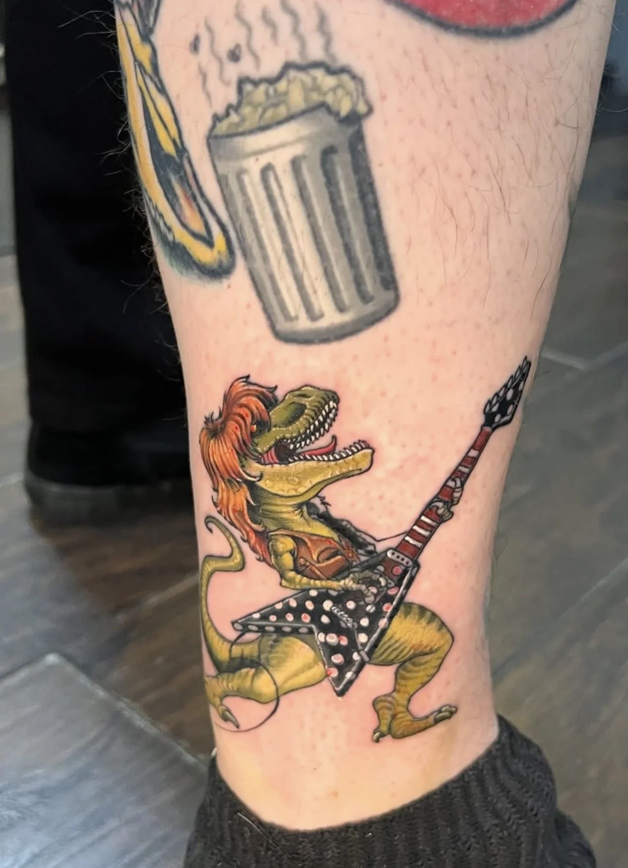 A tattoo of an anthropomorphic dinosaur playing an electric guitar, with a party in the background.