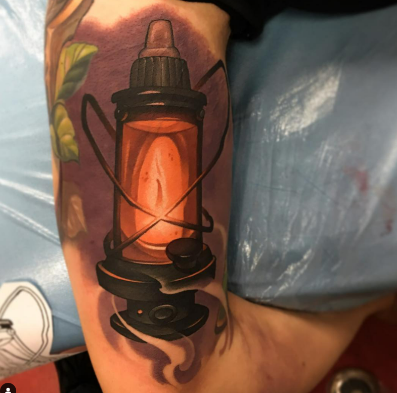 Colorful tattoo of an old-fashioned lantern with a glowing light inside, surrounded by green leaves.