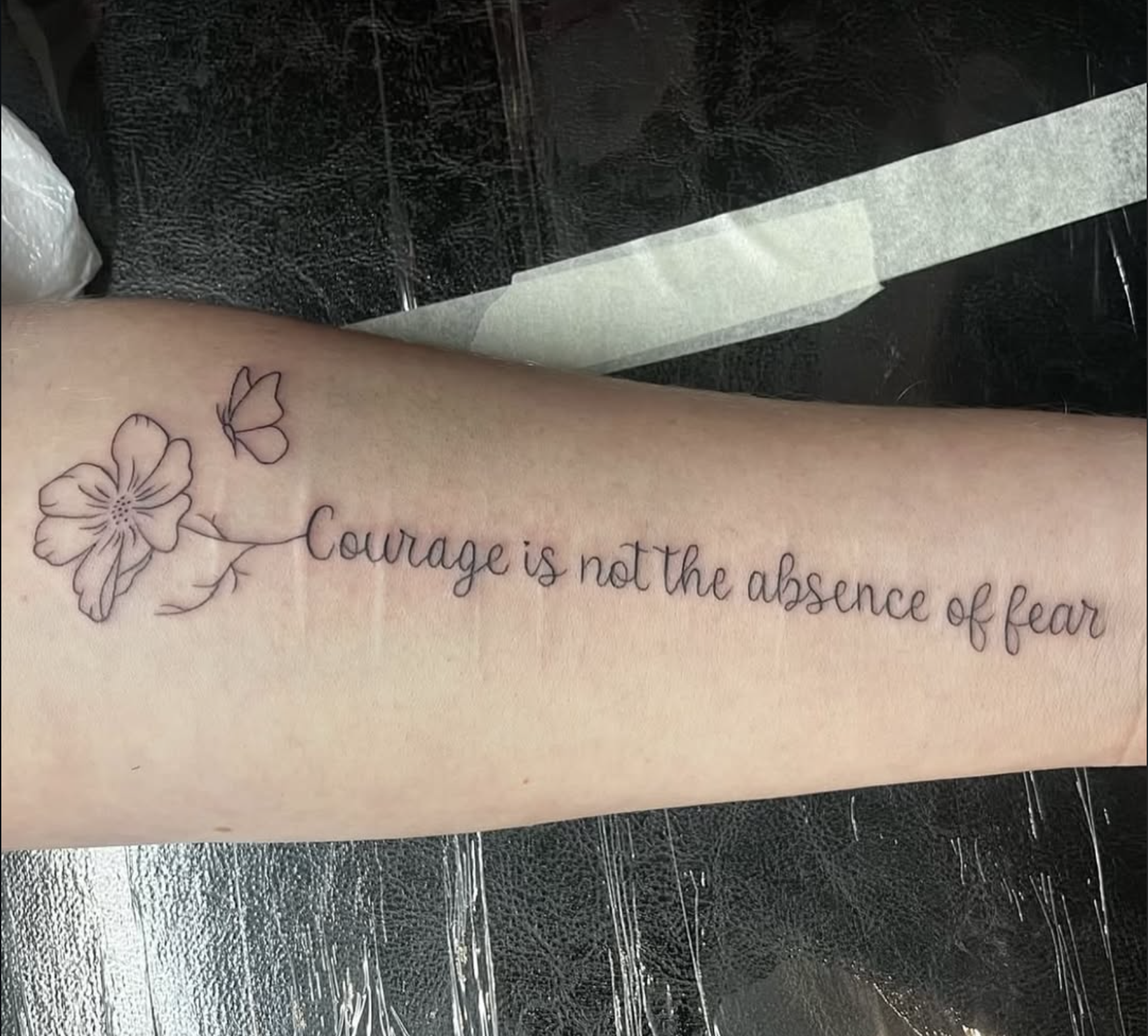 A tattoo on a person's forearm with a flower and two butterflies, accompanied by the phrase 'Courage is not the absence of fear' in cursive writing.