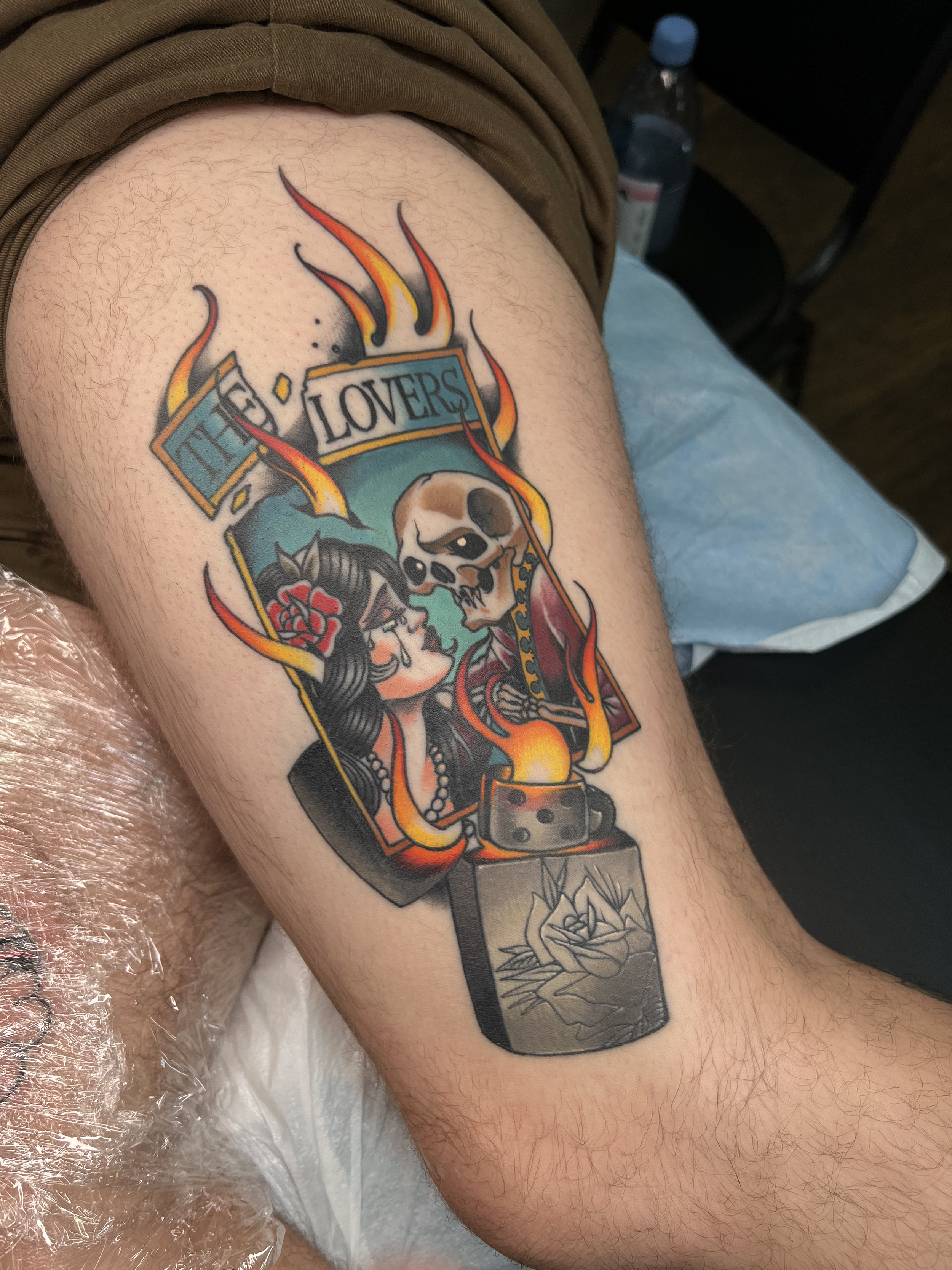 A tattoo on a person's thigh featuring a woman with long black hair, a skull with a rose, and flames, with the words "The Lovers" in a banner.