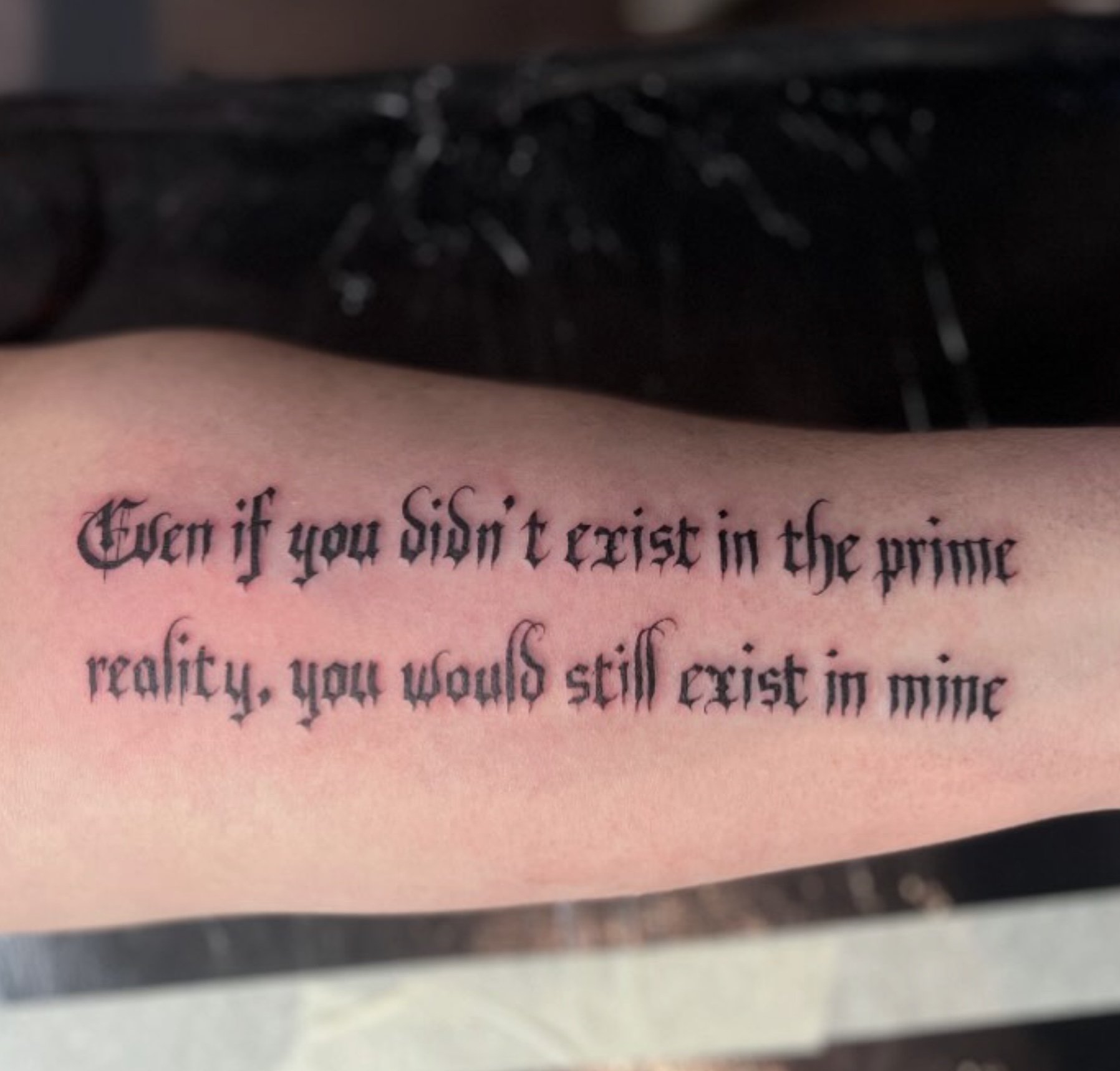 Close-up of a tattooed arm with text in black Gothic font, partially visible.