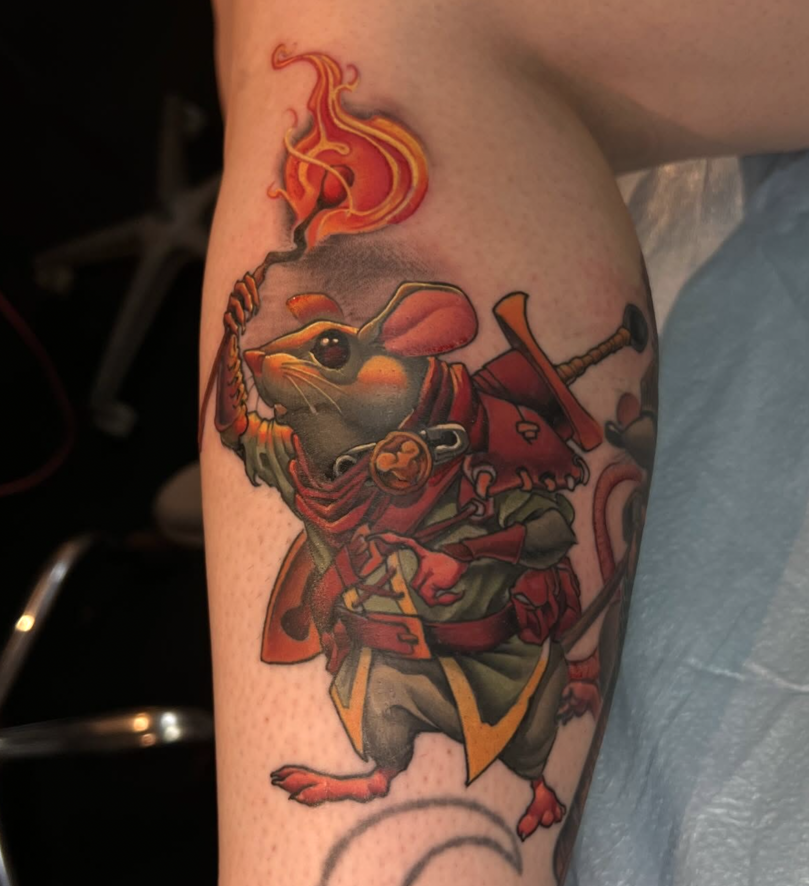 Colorful tattoo of a rat wearing a red cape, holding a sword, with a swirling fire motif above on a person's arm.