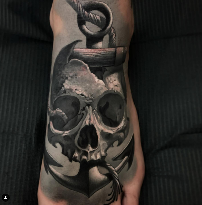 A detailed black and gray tattoo of a skull with a rope tied at the top, and a dagger piercing through the skull, on a person's arm.