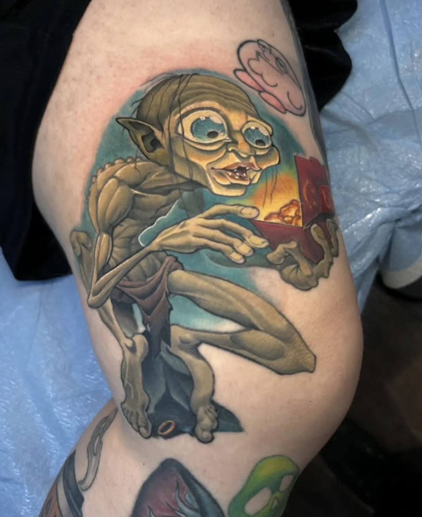 A colorful tattoo of a cartoonish goblin or elf character with large eyes and pointed ears, sitting with one leg up, holding a small red box that is emitting a warm glow. The character has a mischievous expression, and the tattoo includes detailed sh