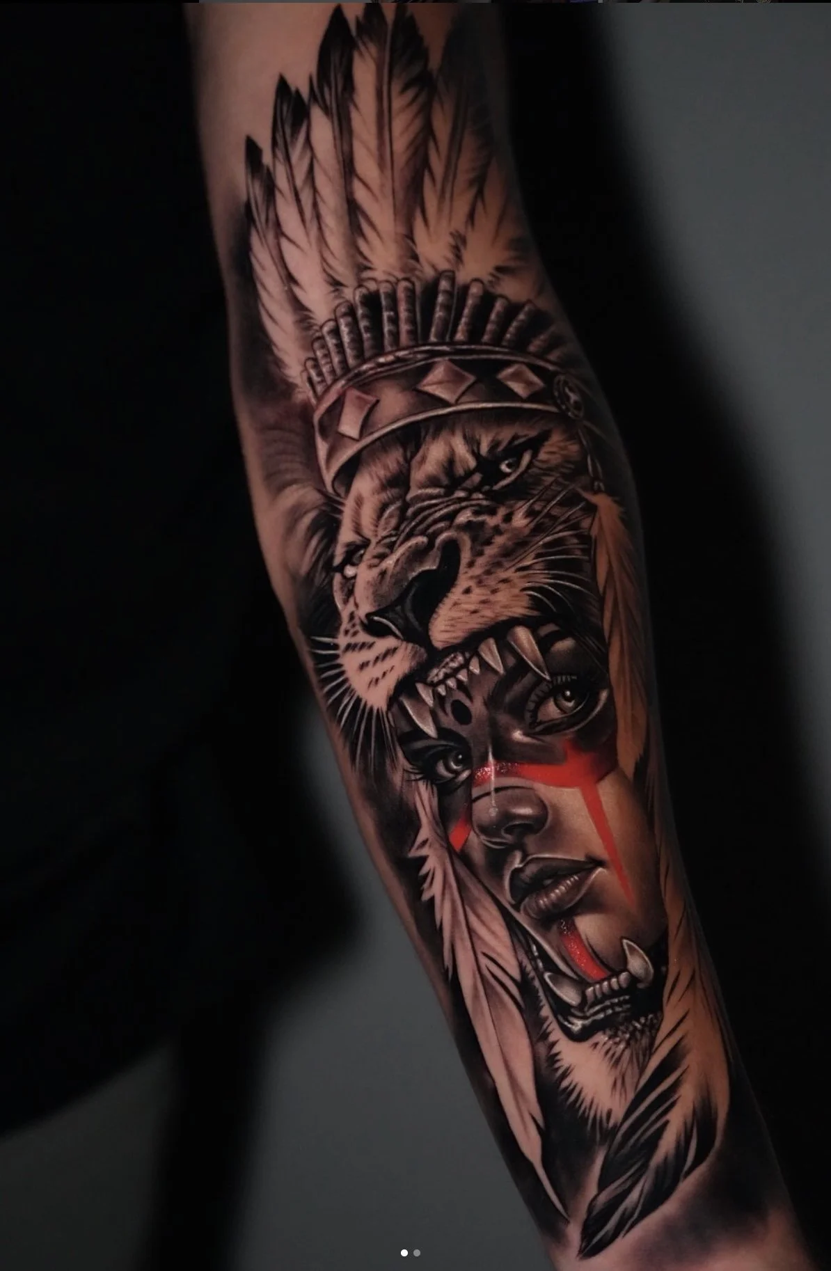 Tattoo of a woman's face with red tribal markings, a fierce tiger wearing a headband, and feathers on the arm.
