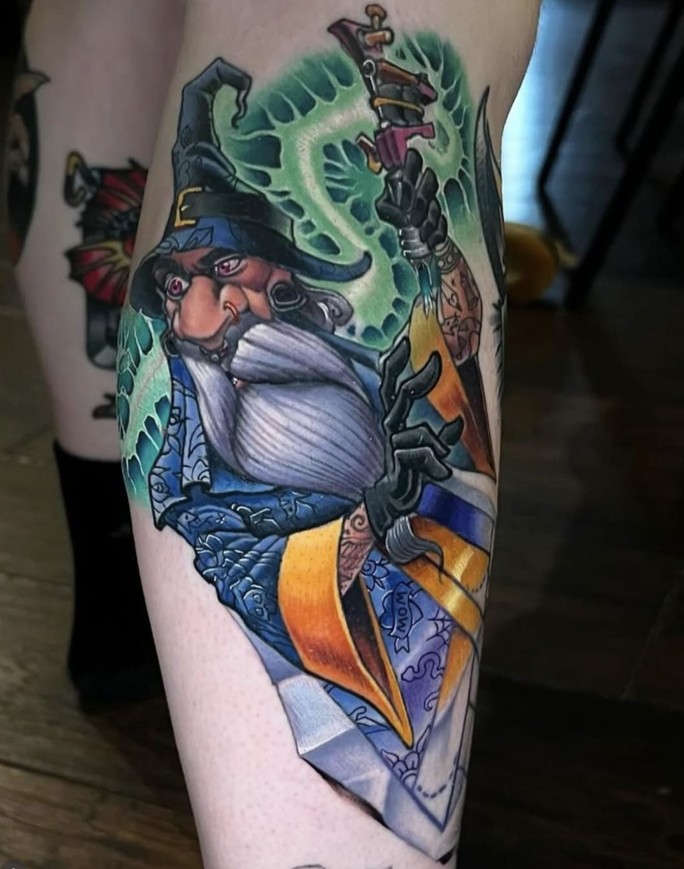 Colorful tattoo of a wizard with a long white beard, wearing a blue hat, holding a sword, with green magical energy swirling around him.