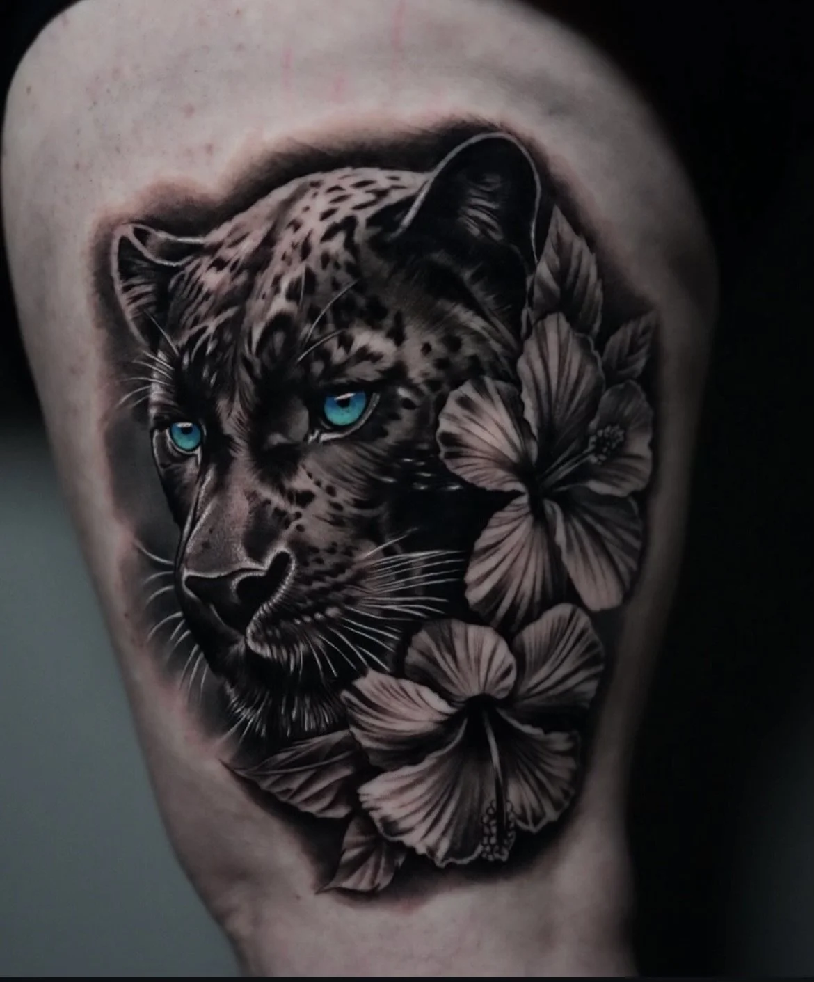 Tattoo of a black panther with vibrant blue eyes surrounded by realistic flowers on a person's shoulder.