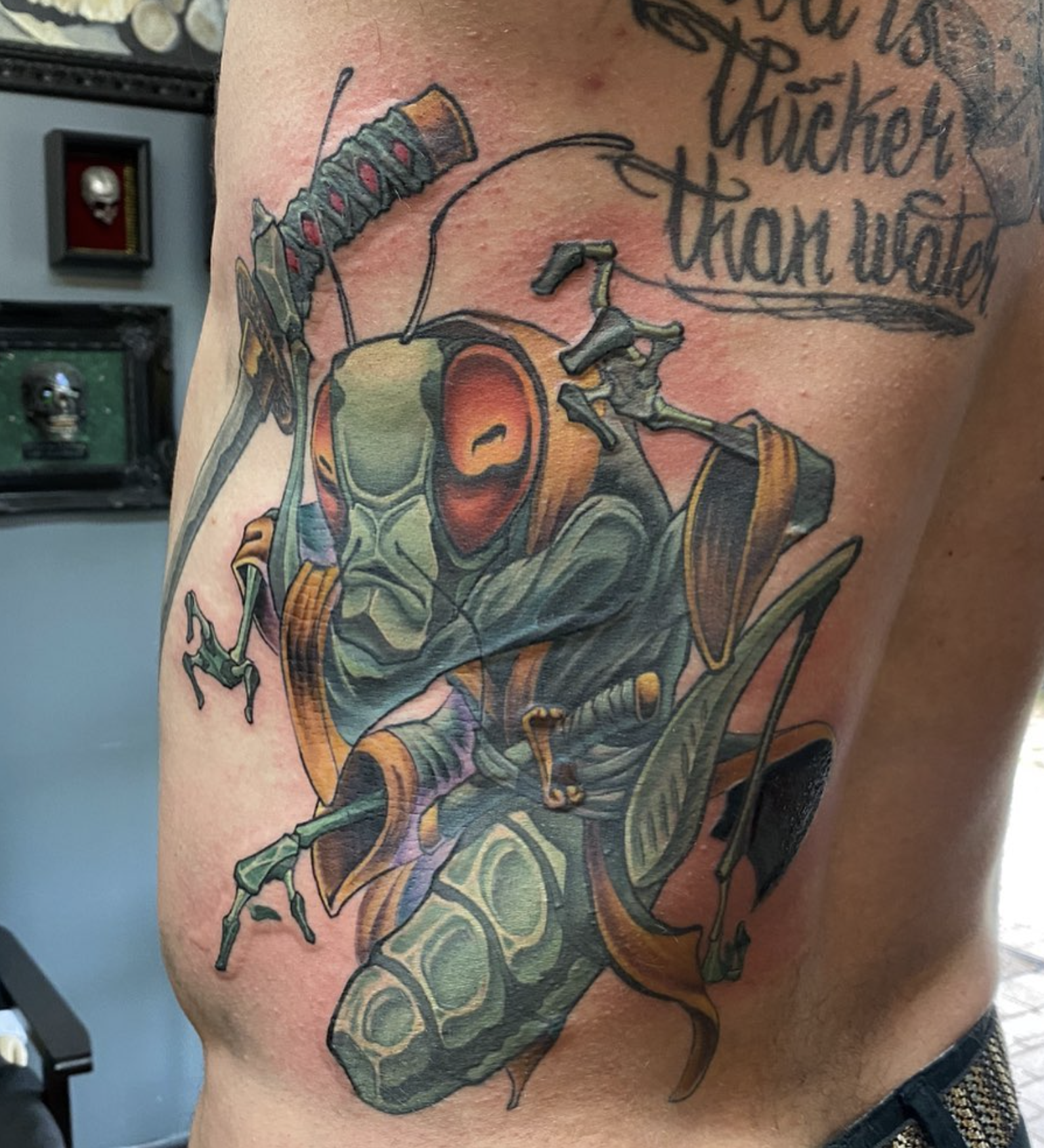 Colorful tattoo of a praying mantis with detailed shading and colors, partly covering some text on the person's arm.