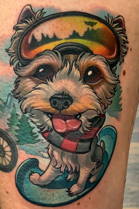 Colorful tattoo of a cute dog with a cheerful expression, wearing a red and black checkered scarf, wearing goggles with a landscape scene, and surrounded by trees and a water spray.