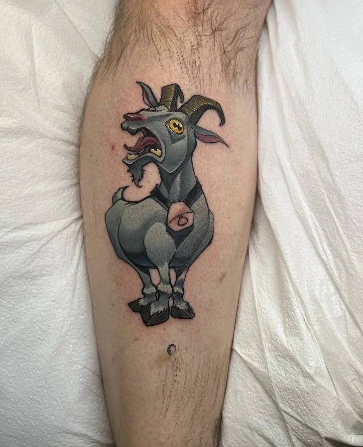Colorful tattoo of a cartoon goat with horns, a beard, and a collar, shown on a person's leg.