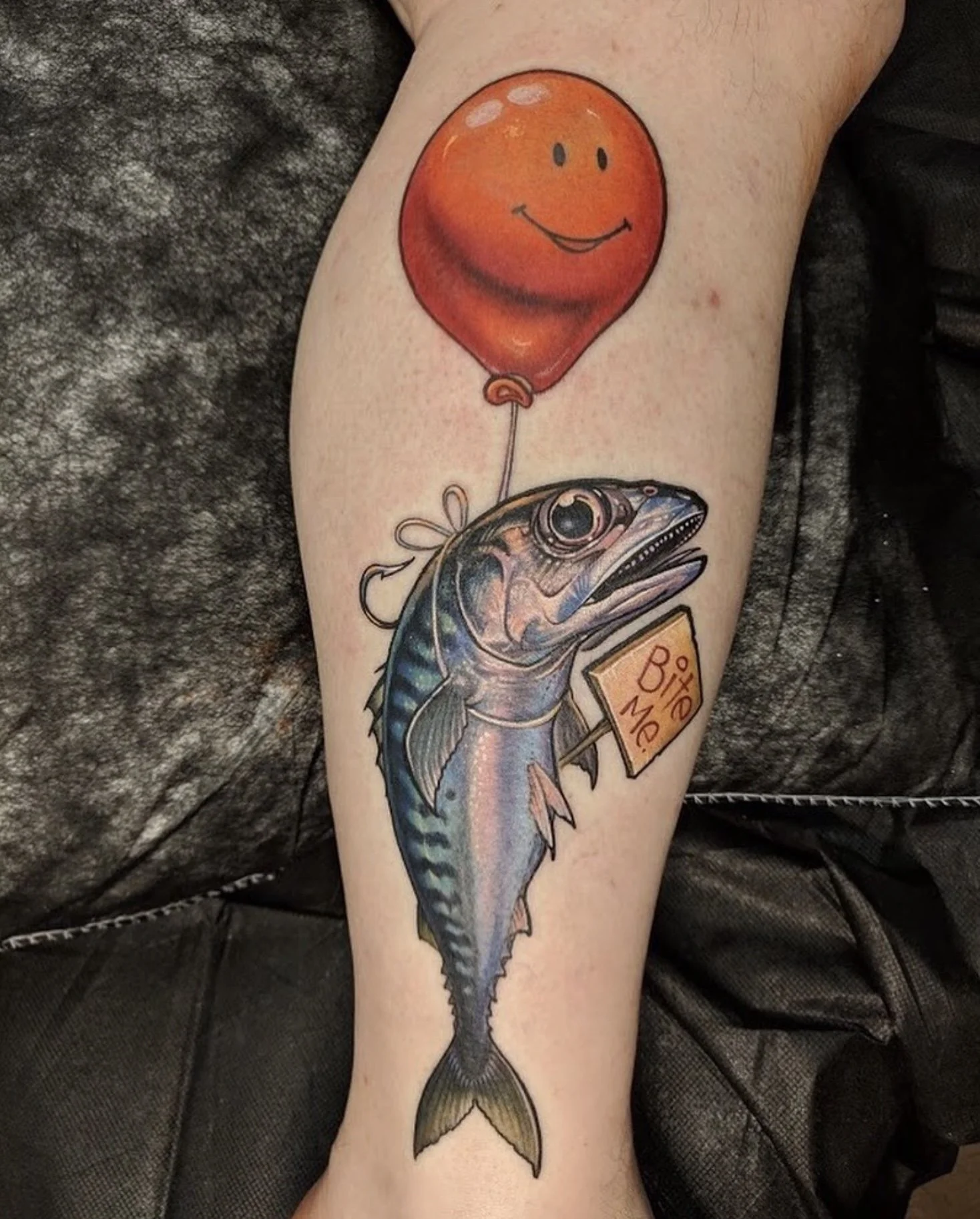 Tattoo of a fish holding a sign that says 'Bite Me' with a red balloon that has a smiling face.
