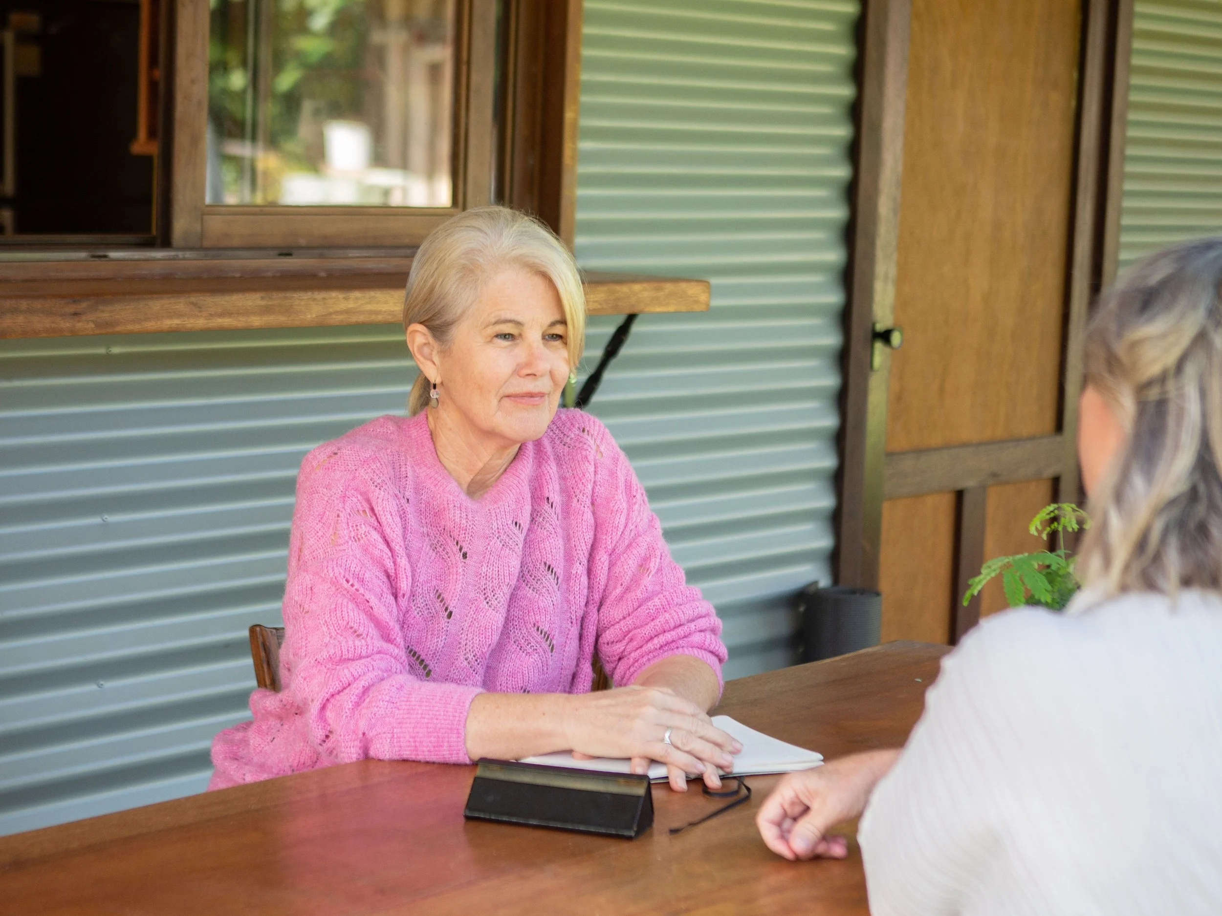 Two women sitting at a wooden table indoors, having a conversation, with one woman wearing a pink sweater and the other a white sweater.