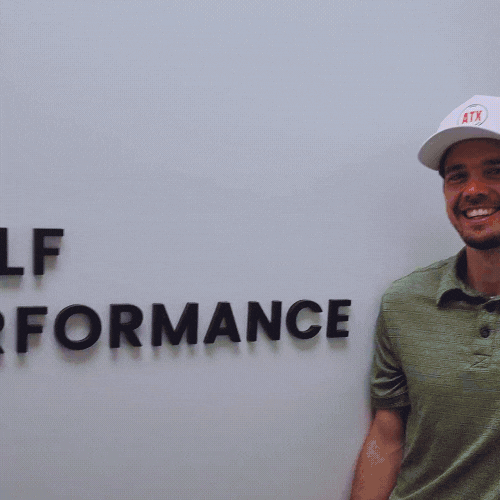 Man smiling in a green shirt and white hat standing next to a partial sign that reads 'PERFORMANCE' on a white wall.