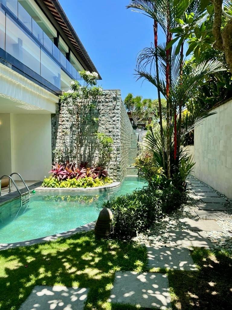 Luxurious pool area with clear water, surrounded by lush tropical plants and modern architecture, featuring stone walls and a sunlit pathway.