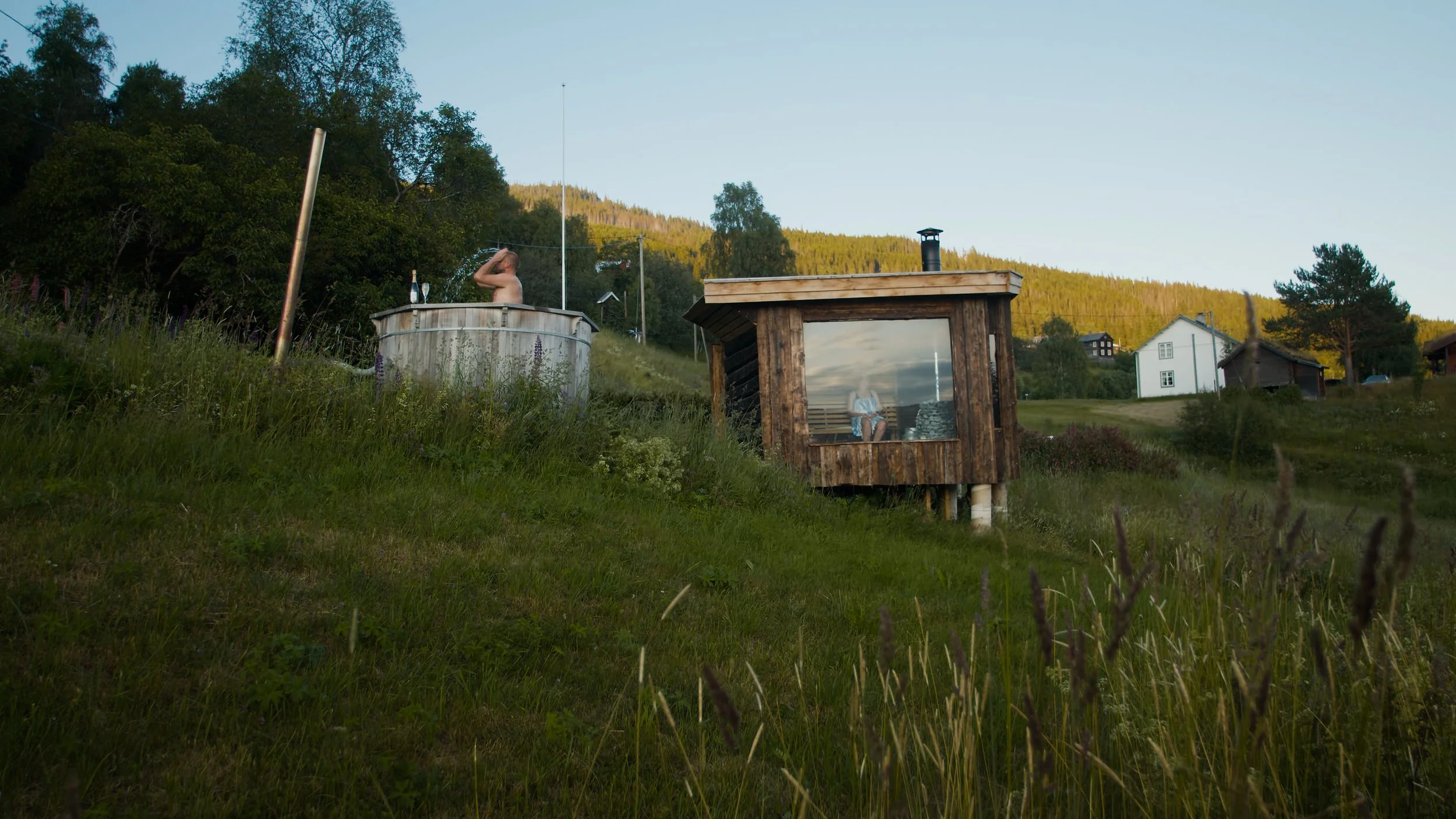 En person i en kaldstamp, omringet av en gresslette med vekster, omgitt av en hage, badstogo og vakker himmel i bakgrunnen.