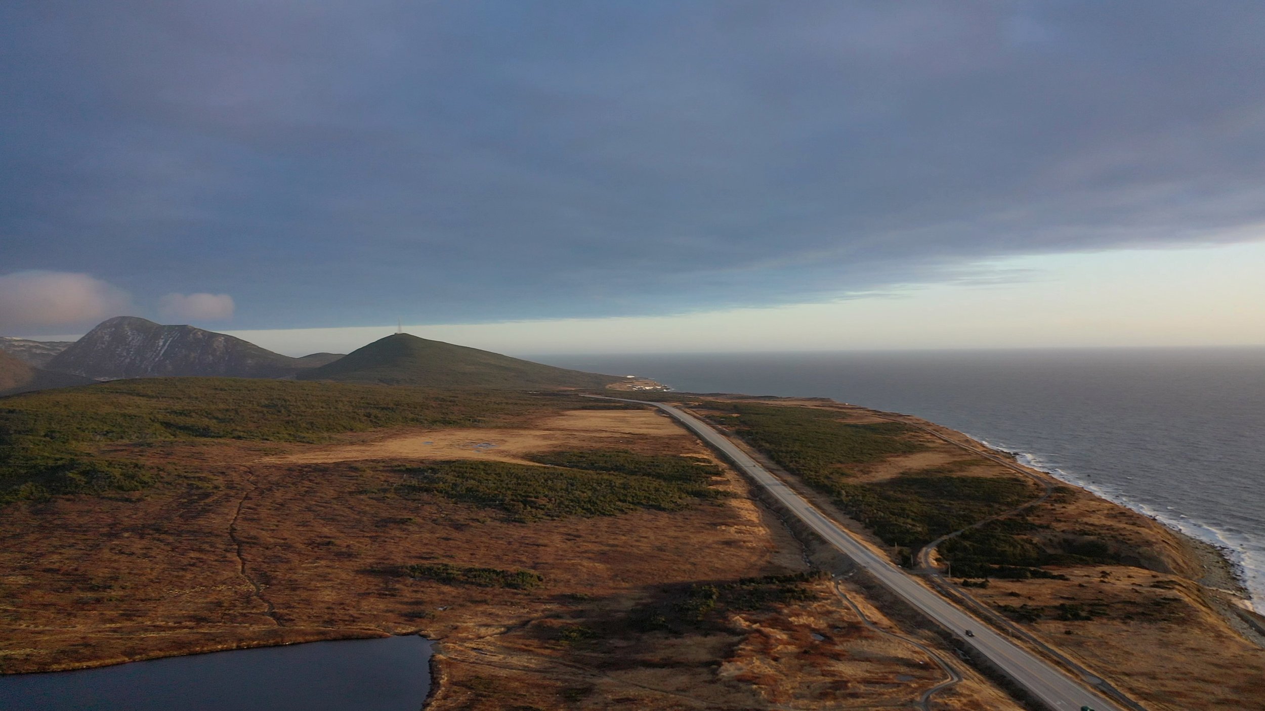 A landscape view of mountains, a road, and ocean under a dark, cloudy sky.