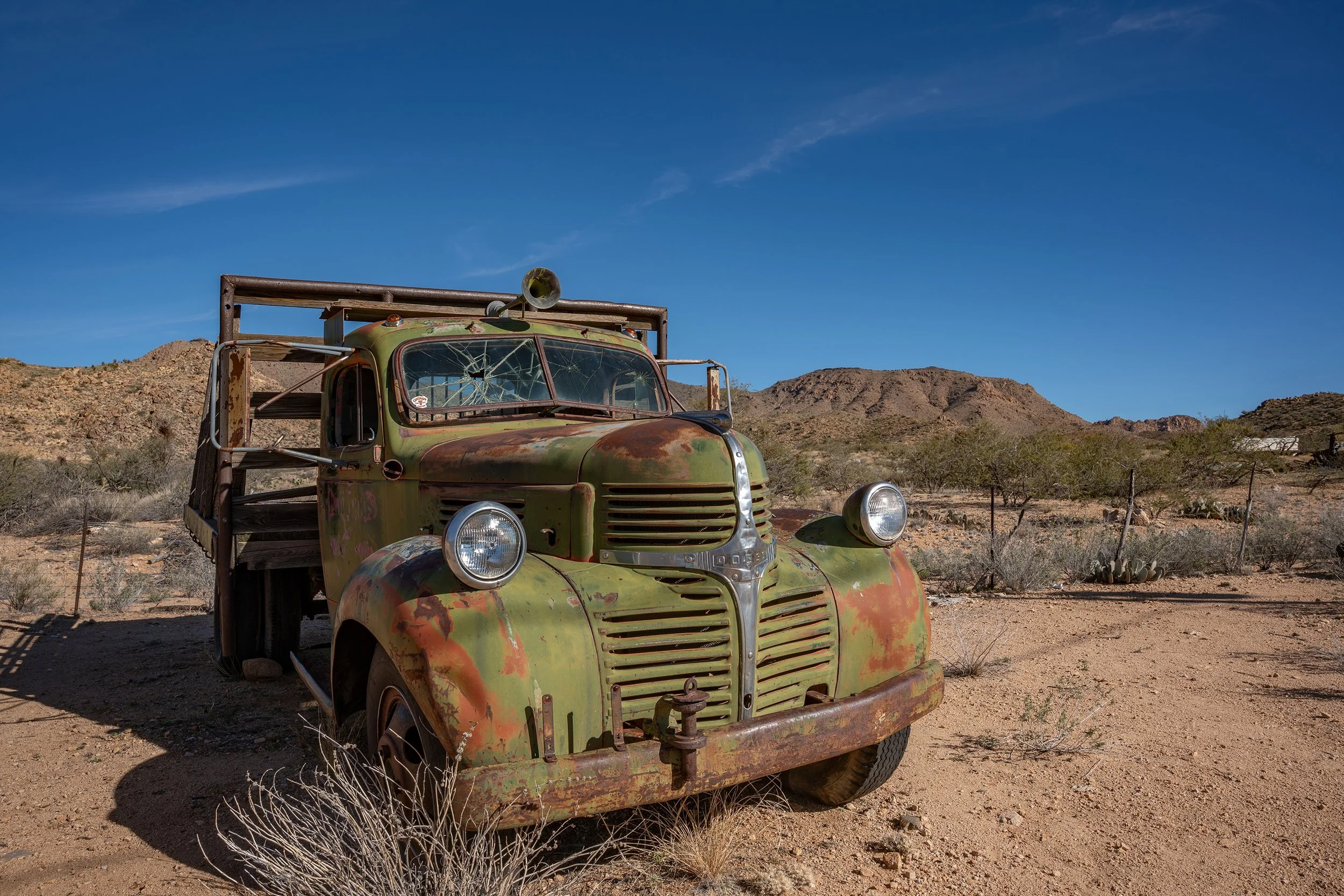 Old rusty green truck in a desert landscape with mountains and clear blue sky in the background.