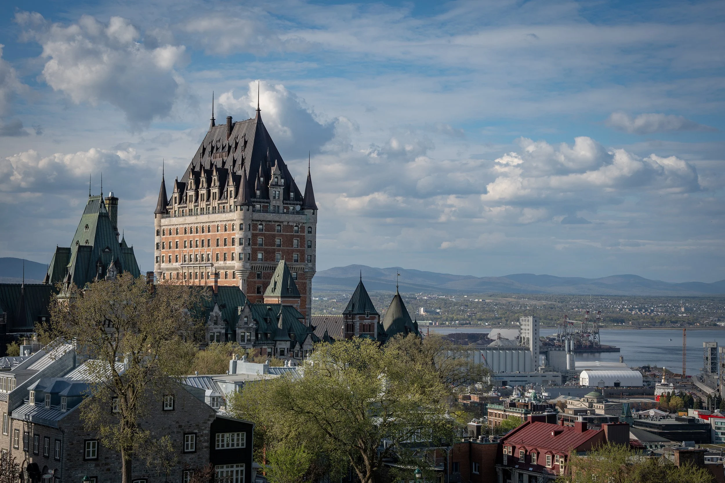 A cityscape featuring a prominent castle-like building with tall, pointed rooftops, a river in the background, and blue sky with scattered clouds.