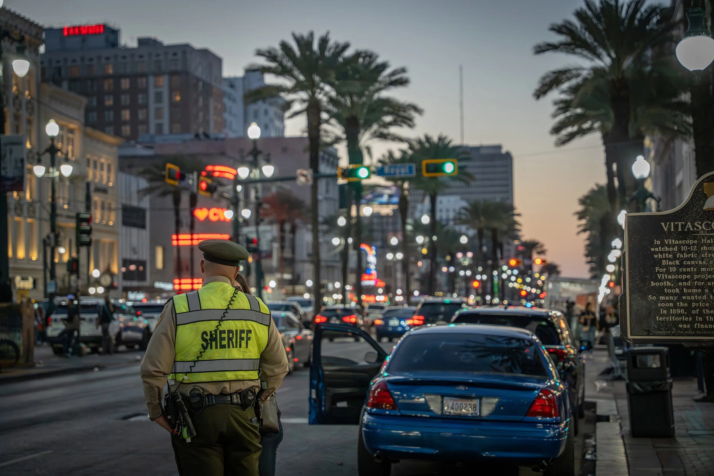A city street at dusk with a sheriff officer standing with his back facing the camera, wearing a yellow reflective vest that says 'SHERIFF'. There are cars on the street, palm trees, and illuminated signs in the background. New Orleans