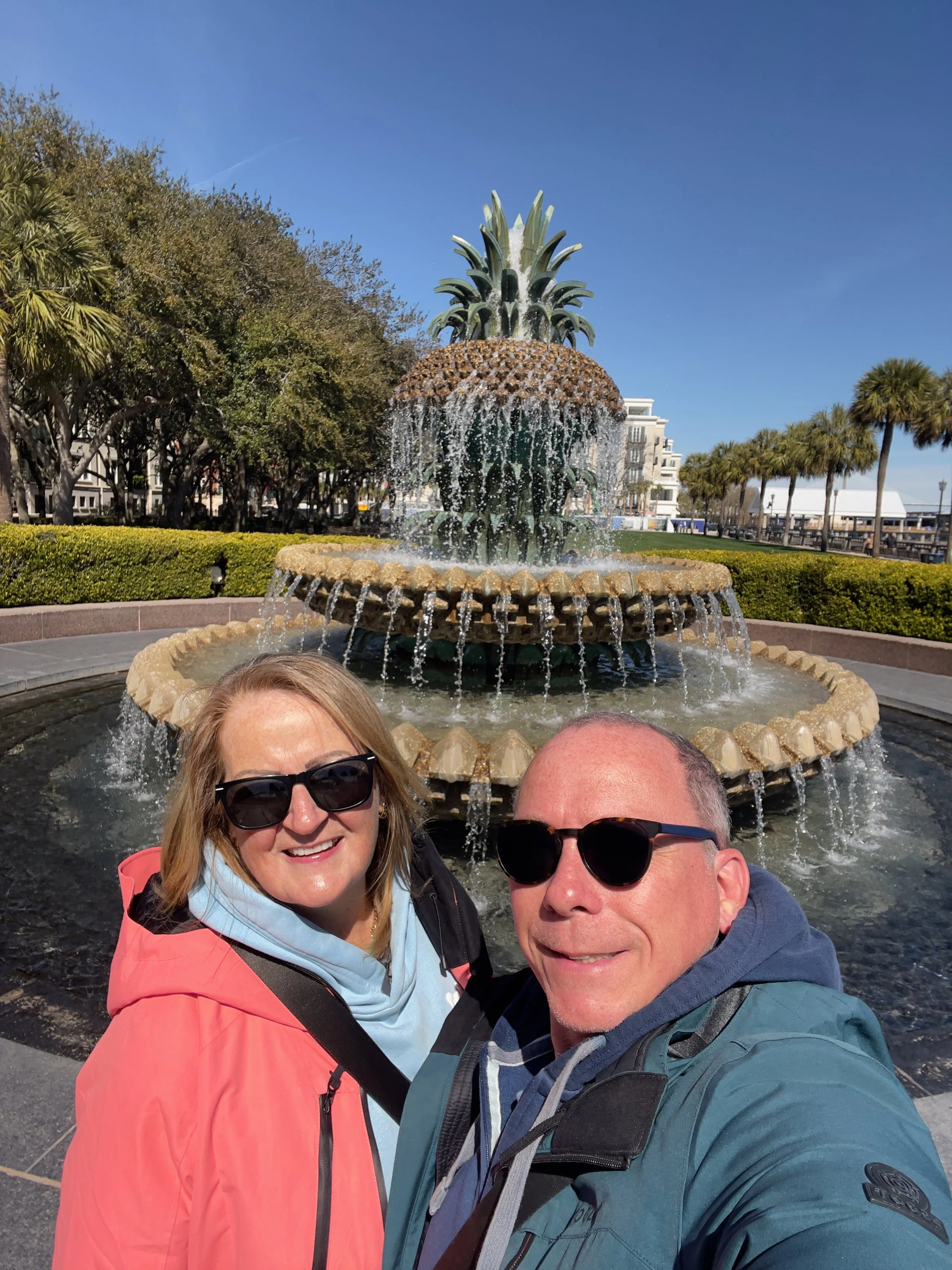 Two people taking a selfie in front of a pineapple-shaped fountain with water cascading down, trees, buildings, and a clear blue sky in the background.