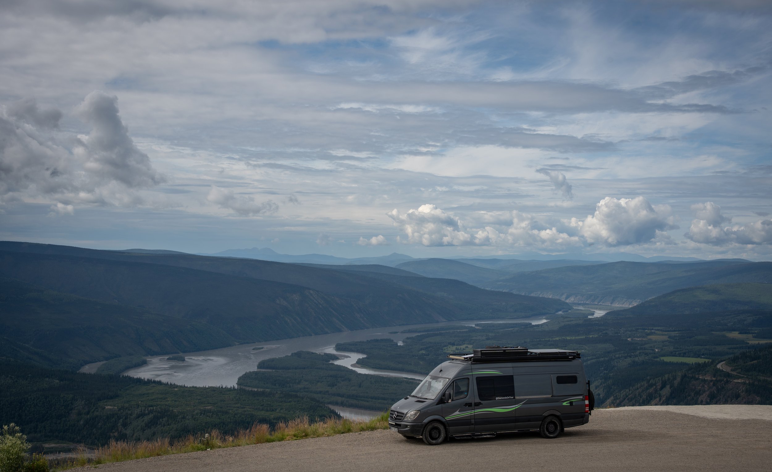 A grey camper van parked on a gravel viewpoint overlooking a river winding through lush green mountains under a cloudy sky.