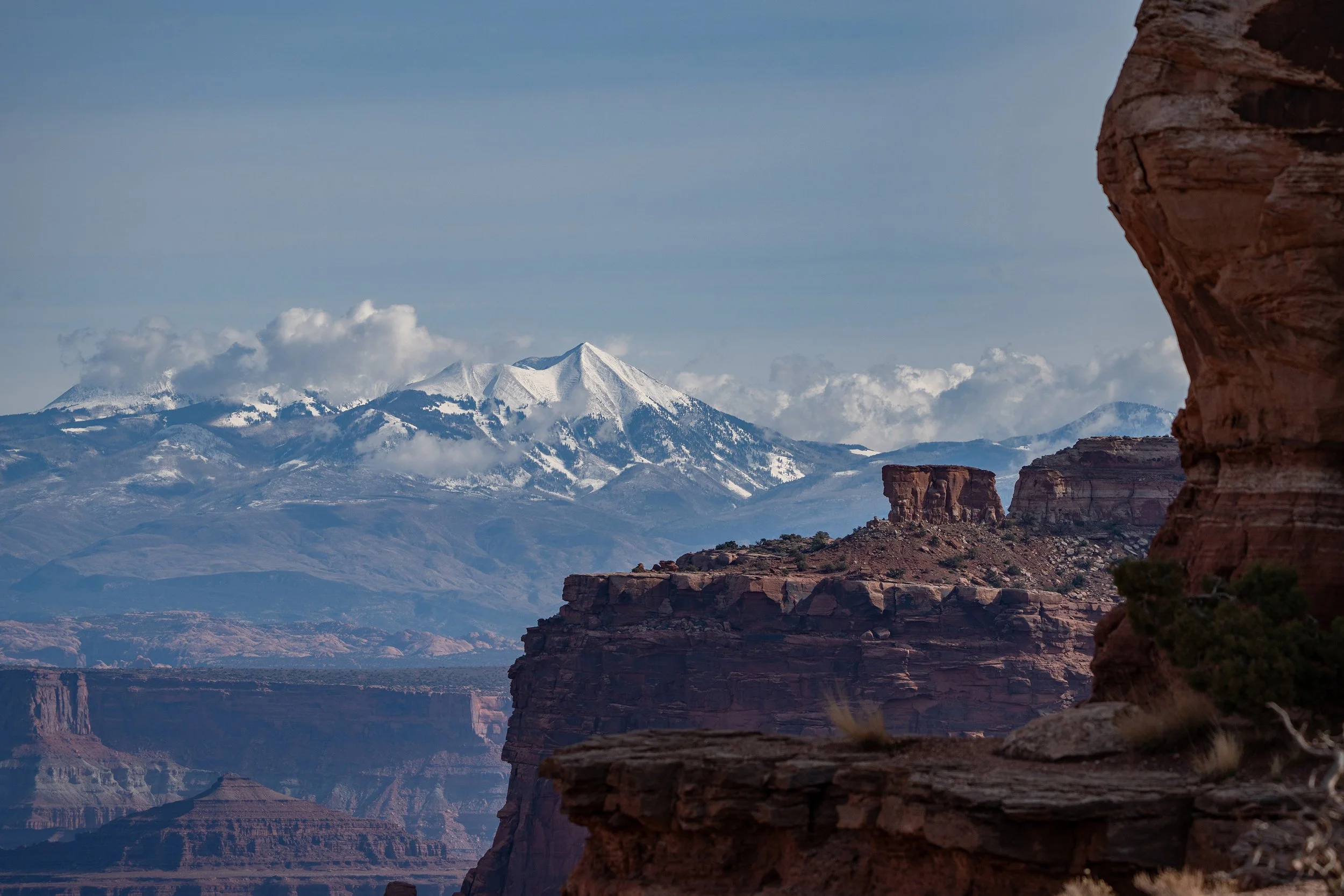 Snow-capped mountains in the background with reddish rock formations and cliffs in the foreground, clear blue sky with some clouds.
