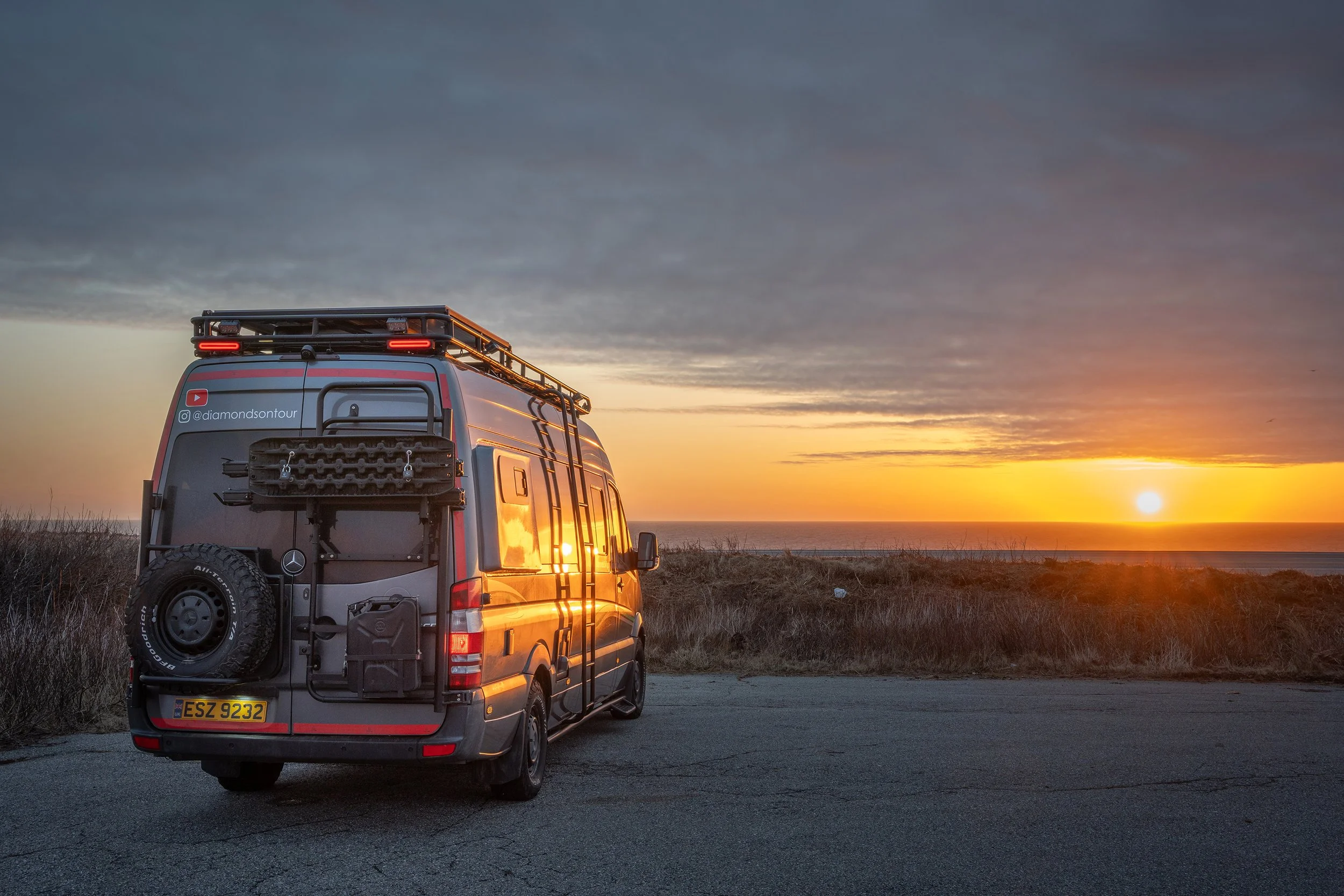 A camper van parked on a scenic overlook at sunset, with a cloudy sky and the ocean in the background.