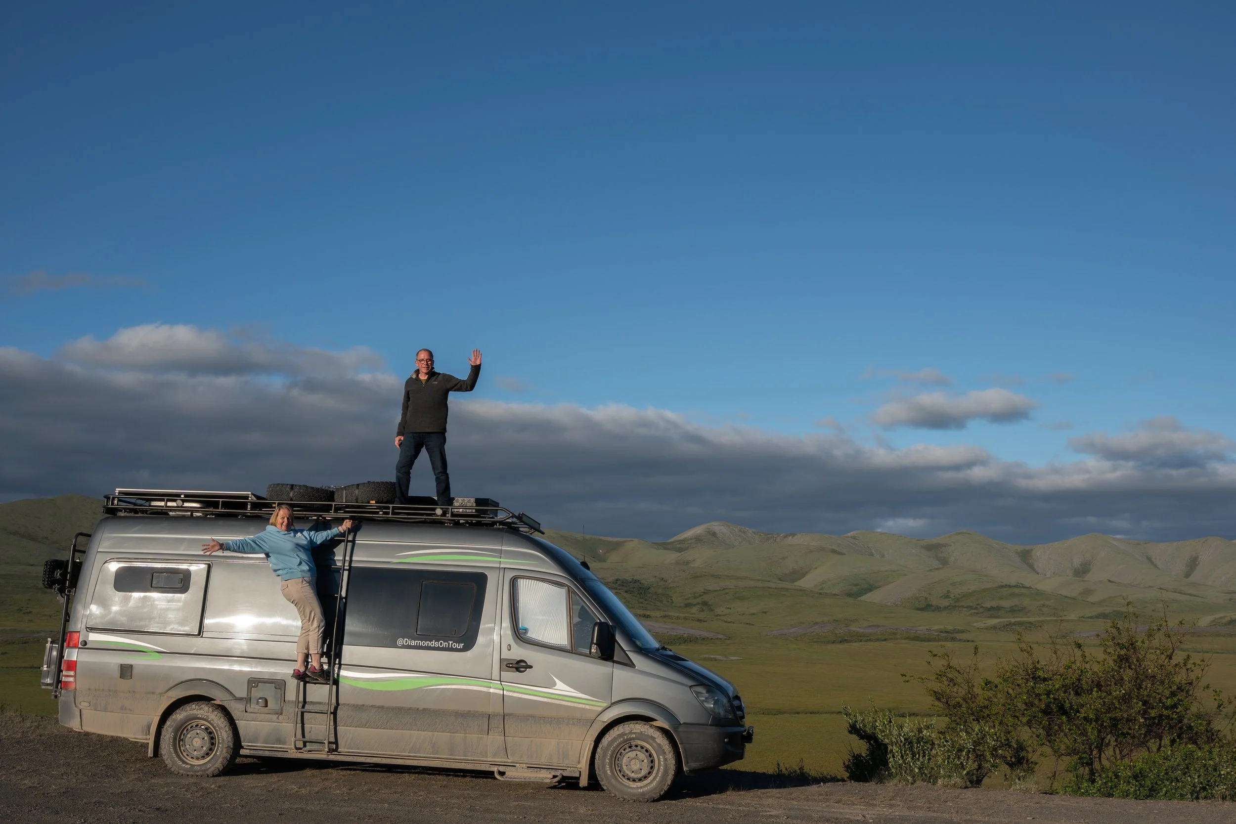 Two people are posing outside a van in a scenic landscape with rolling green hills and a blue sky with scattered clouds. One is standing on the roof of the van, waving, while the other is hanging on to the ladder, extending her arms.