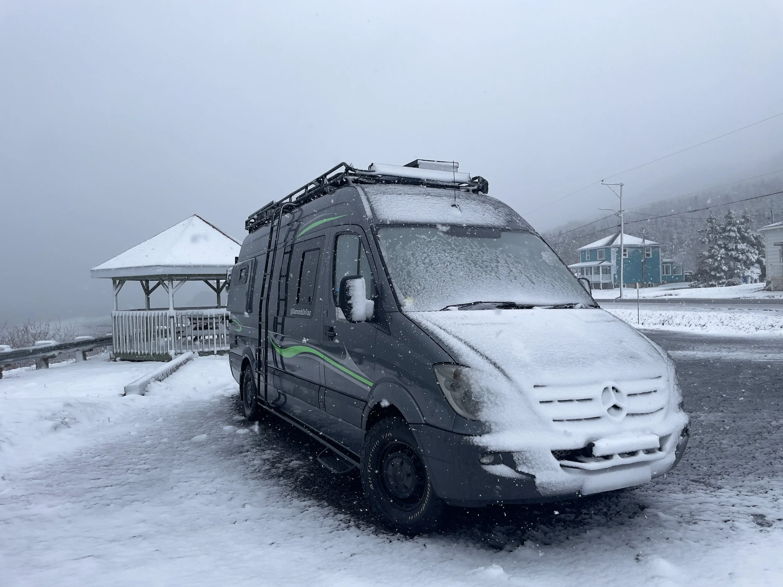 Snow-covered van parked in a snowy yard near houses, with snow on the ground and trees, and a gray, overcast sky.