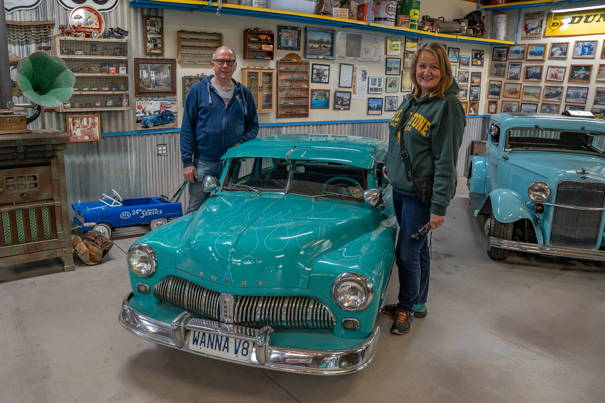 Two people, a man and a woman, standing inside a vintage car museum. The man is wearing glasses and a blue hoodie, and the woman is wearing a green hoodie, both smiling. In front of them is a small, turquoise vintage car with a racing stripe design. 