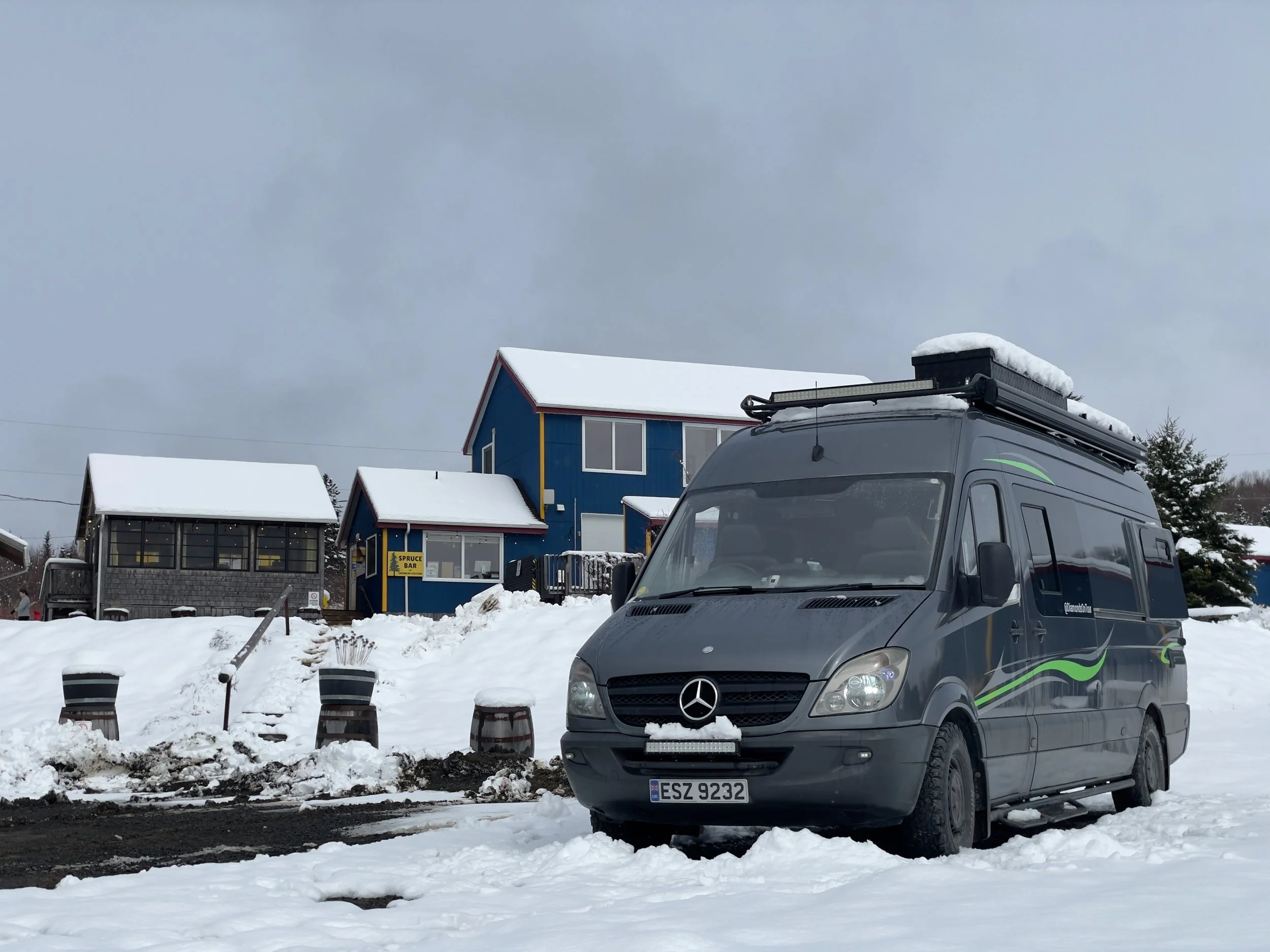 Snow-covered parking lot with a black Mercedes-Benz van in the foreground, colorful houses in the background, and snow on the ground and roofs.