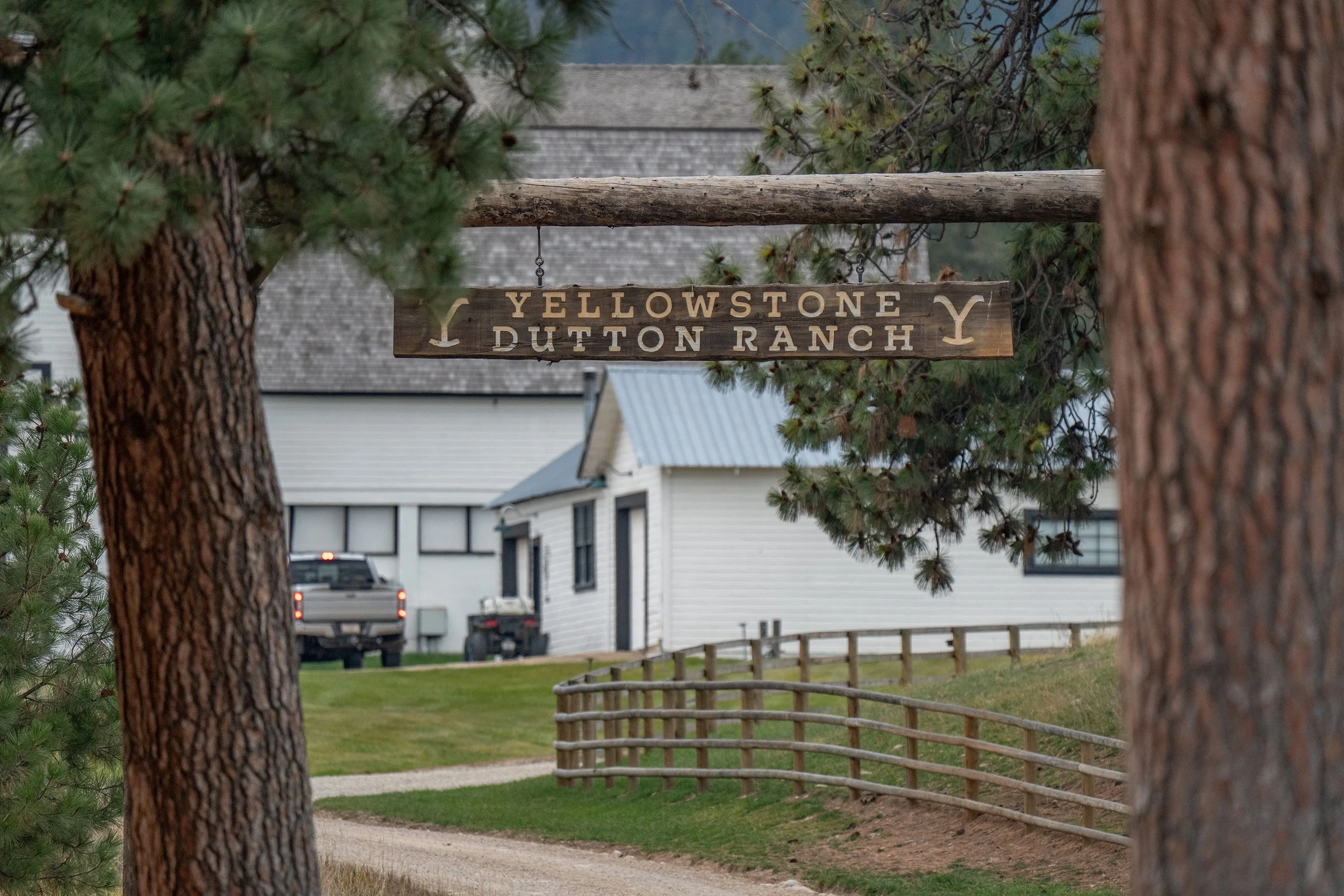 A wooden sign hanging between two pine trees reads "Yellowstone Dutton Ranch" with cowboy hat and Y symbols, in a rural area with houses, a gravel path, and a wooden fence in the background.