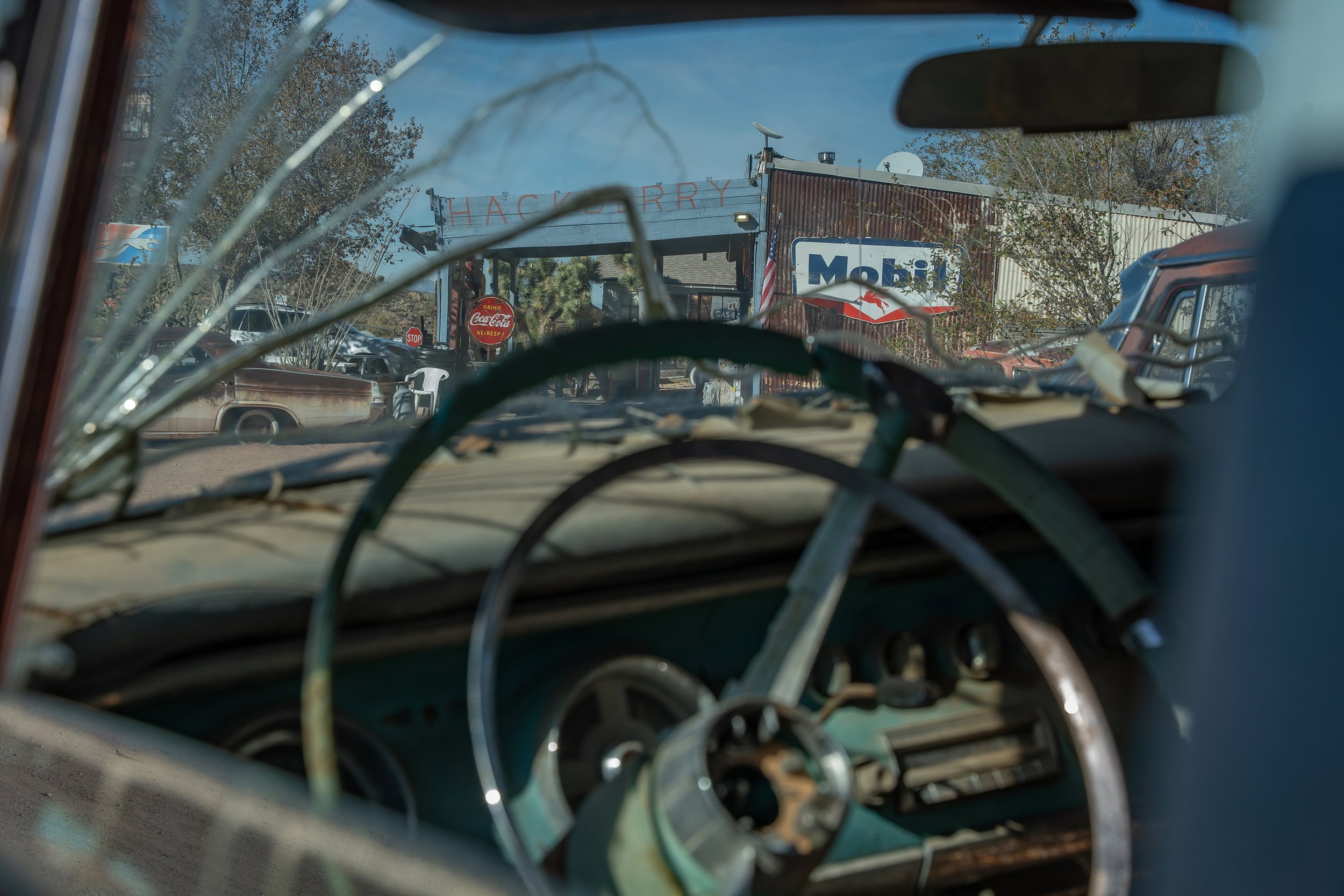 The image shows a view from inside an old, rusted vehicle looking out through the windshield with a broken windshield. In the background, there is a small restaurant or shop with a sign that reads 'HACKBERRY' and a 'MOBILE' sign, along with trees and