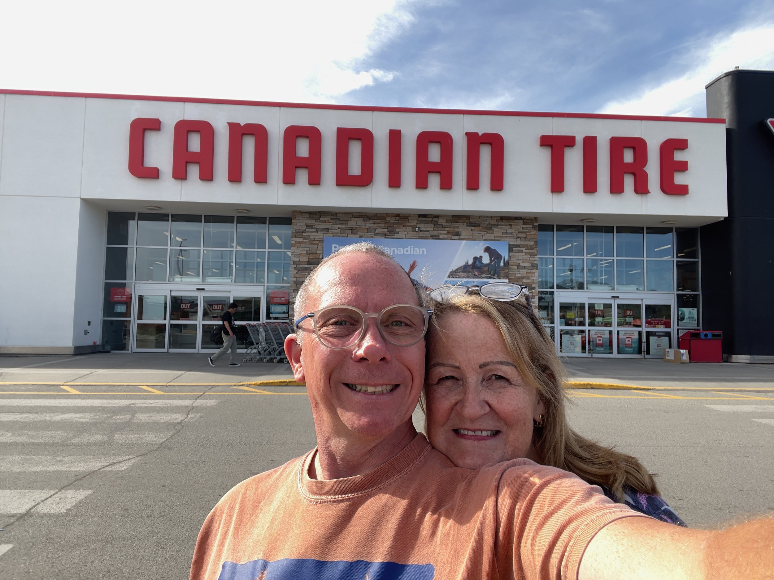 A selfie of an older man and woman standing outdoors in front of a Canadian Tire store, with the store's large sign visible above their heads.