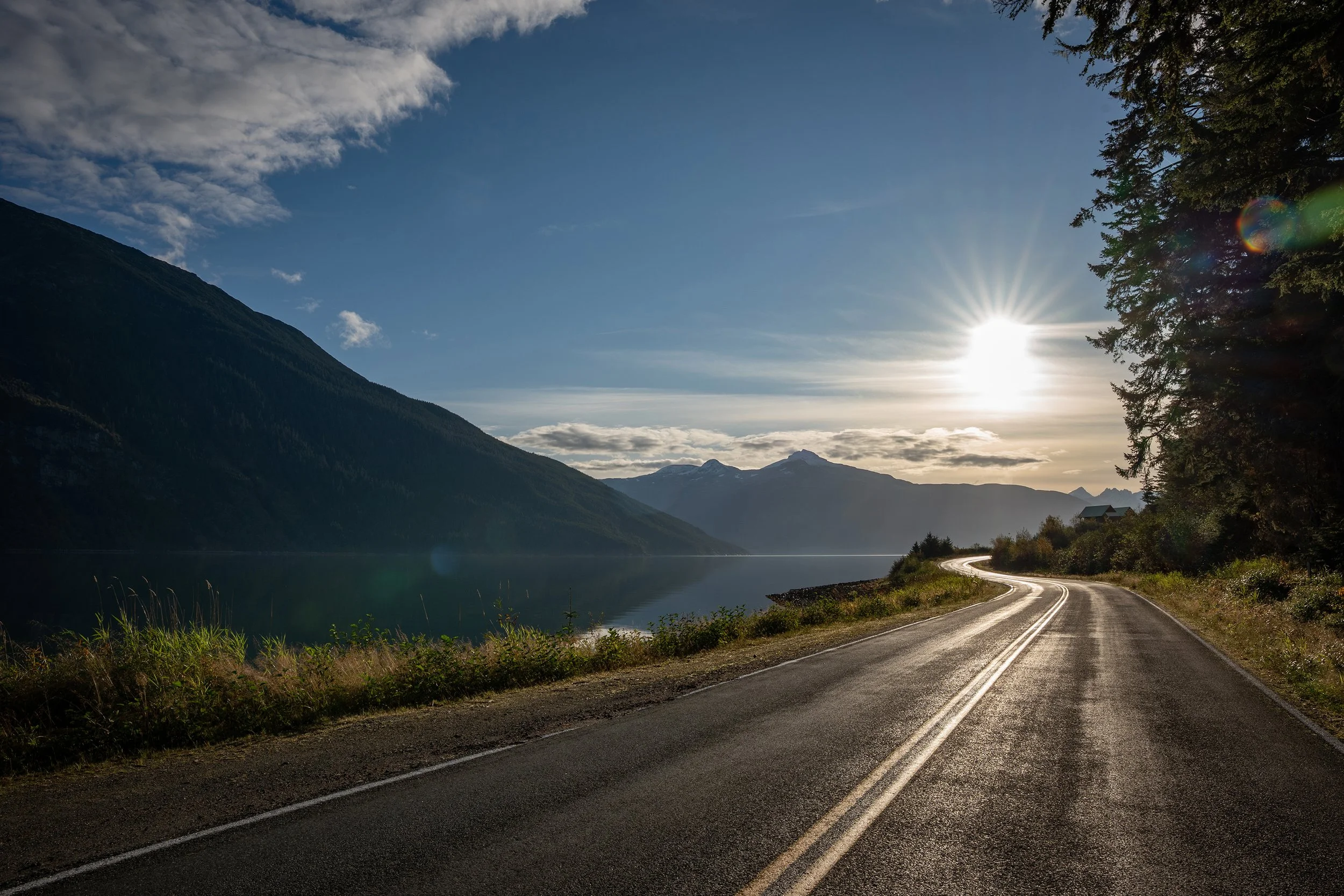 A winding road runs alongside a lake in a mountainous landscape. The sun is shining and partially obscured by trees, casting a bright flare. The sky is mostly clear with some clouds. Haines Alaska.