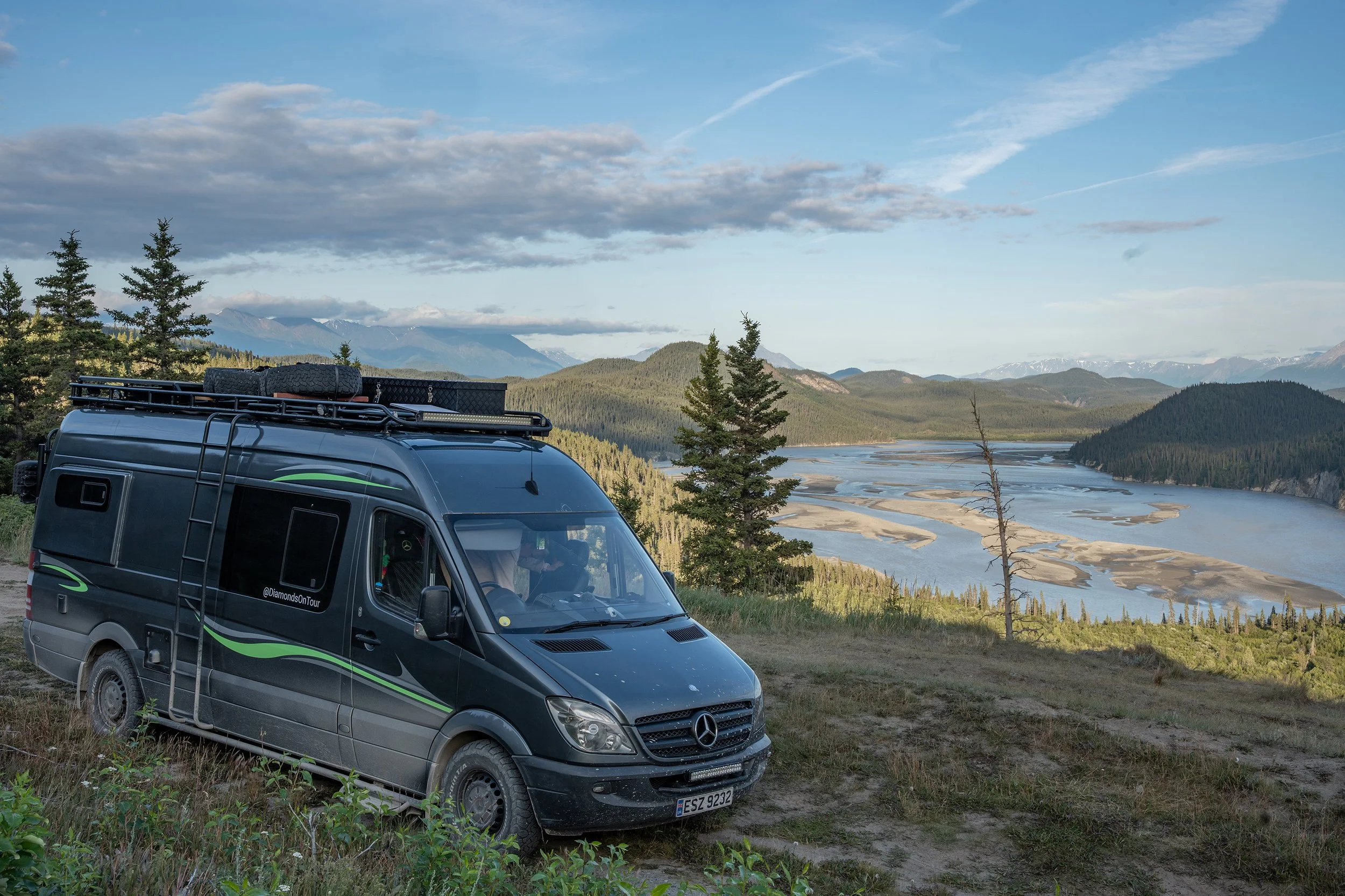 A black camper van with a roof rack and spare tire parked on a grassy area overlooking a river valley with mountains in the background under a partly cloudy sky.